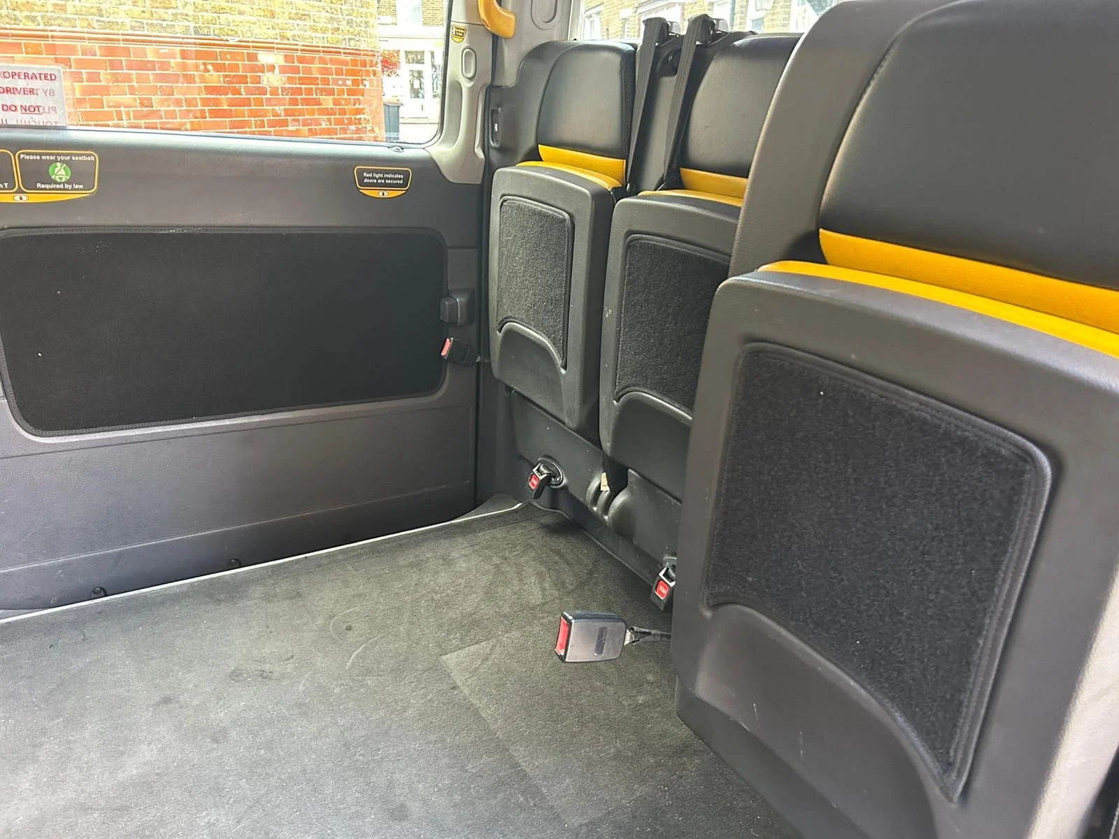 Interior of a passenger van with empty foldable seats, seatbelt latches, and windows showing a brick building outside.