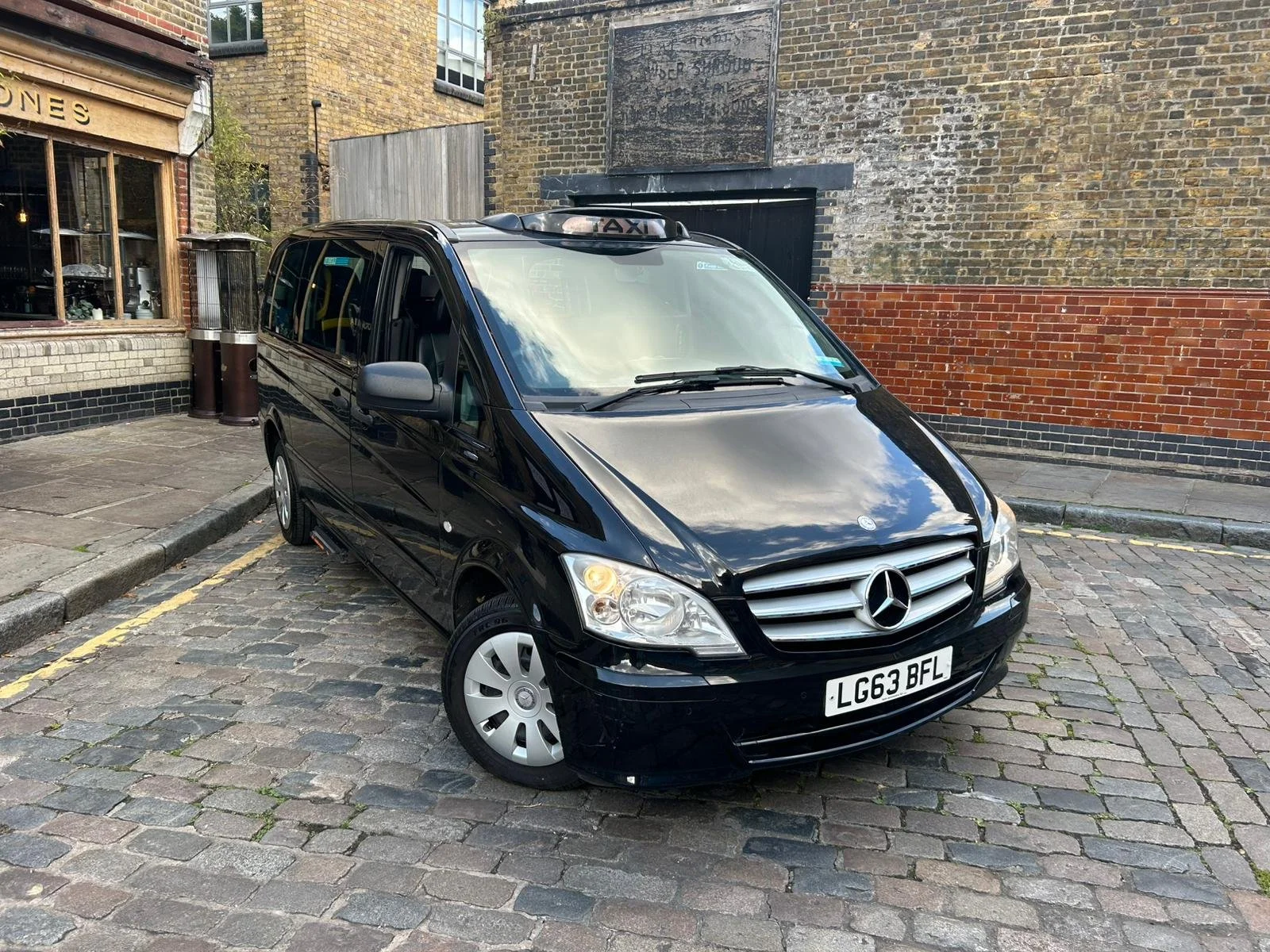 Black Mercedes-Benz taxi van parked on cobblestone street in an urban area with brick and stone buildings in the background.