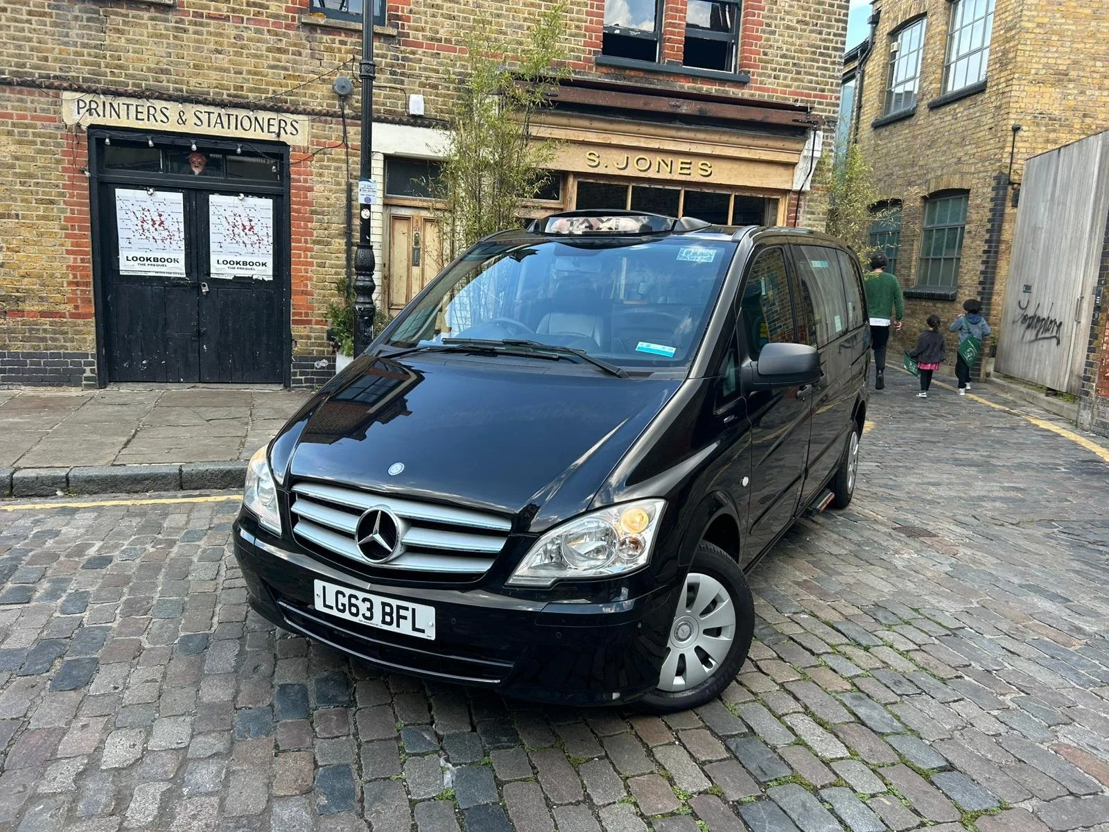 Black Mercedes-Benz van parked on a cobblestone street near brick buildings, with people walking in the background.