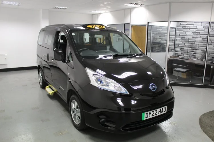 Black Nissan taxi van with a yellow taxi sign on top, parked indoors in a showroom or garage, with a license plate reading DY22 HAU.