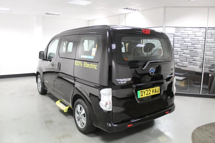 Black electric van with yellow step and '100% Electric' written on the side, parked indoors near glass wall with offices inside.