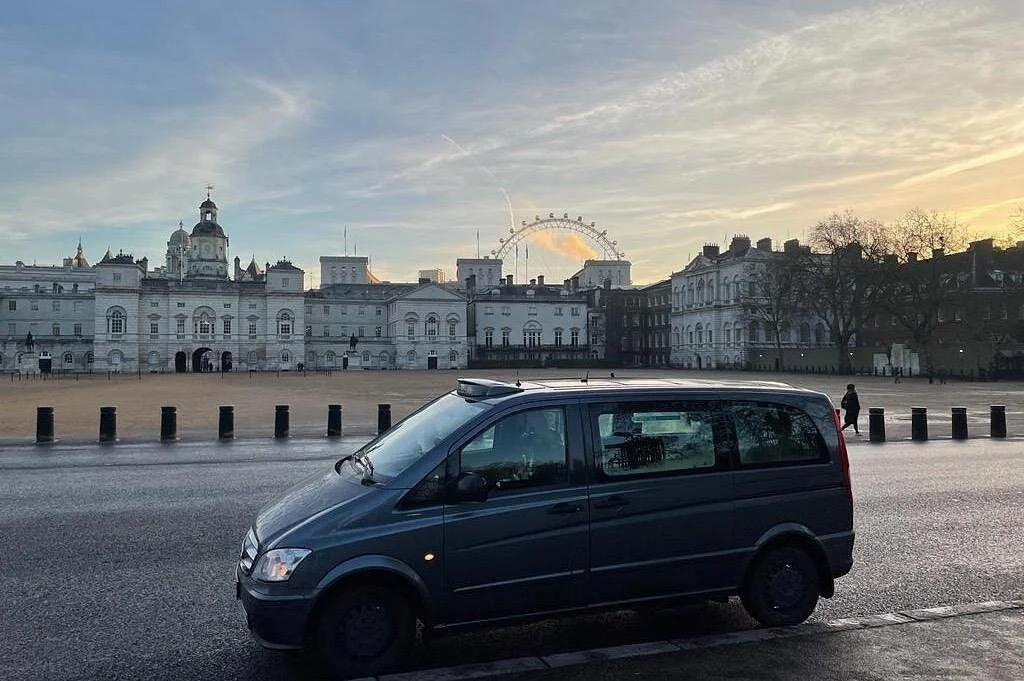 A black van parked on the side of a city street with historic buildings and a Ferris wheel in the background at sunset.