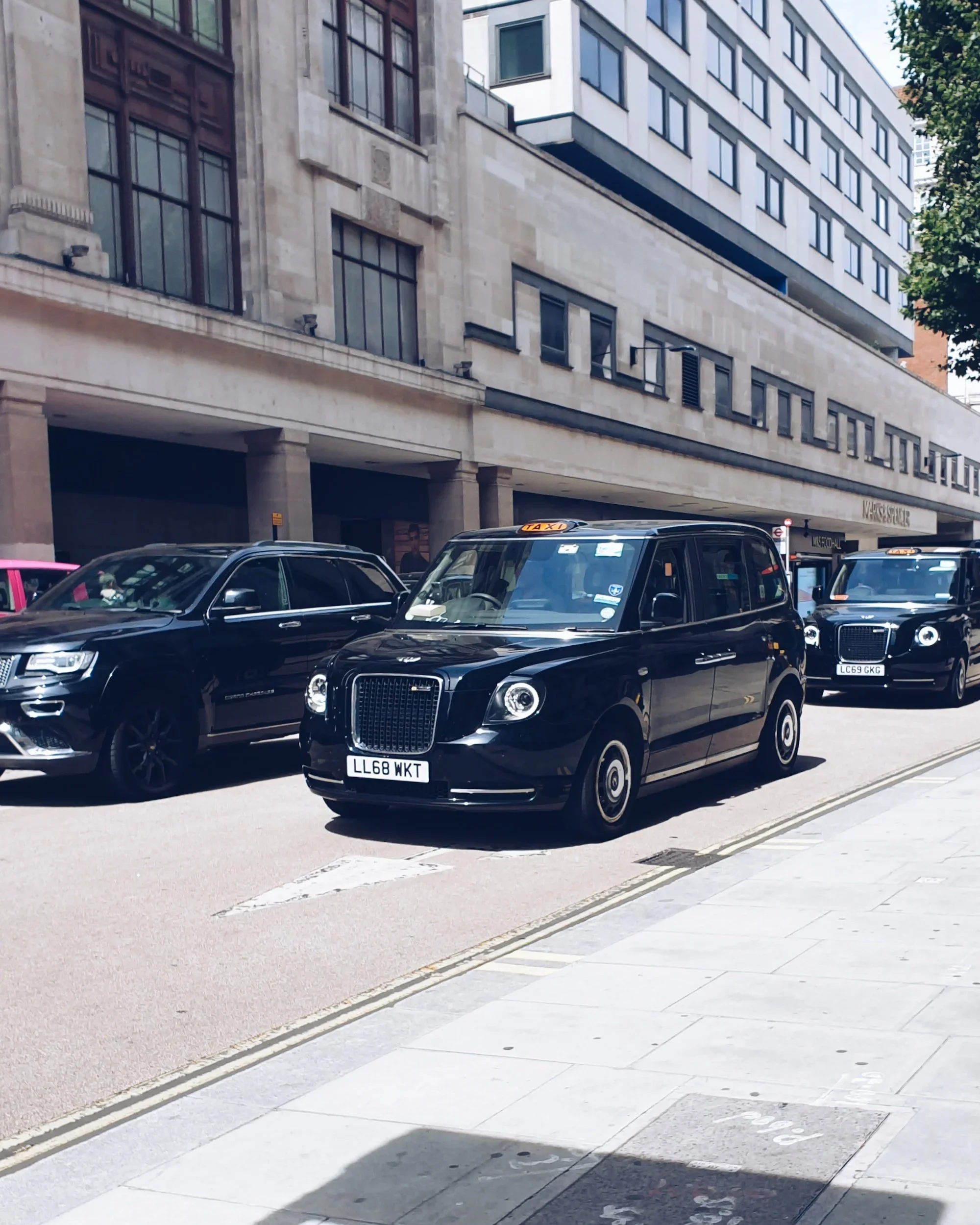 A black taxi cab parked on the street in front of a large building with beige stone walls. Other luxury cars are also parked nearby.