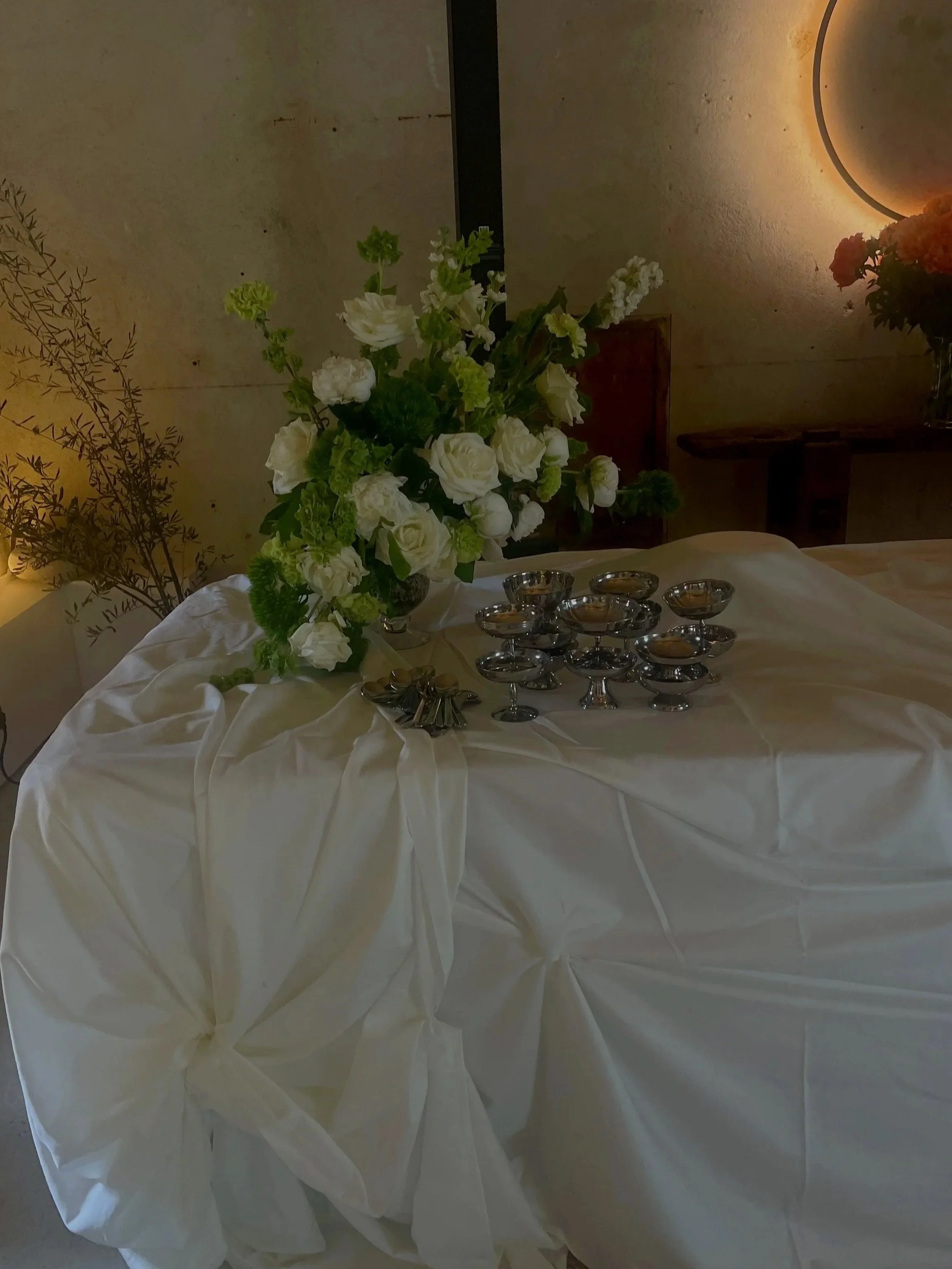Table recouvert d'une nappe blanche avec un arrangement floral composé de roses blanches, et plusieurs petits bols en métal. En arrière-plan, un mur beige avec un lampadaire circulaire émettant une lumière chaleureuse, et une autre fleur en pot visible sur le côté.