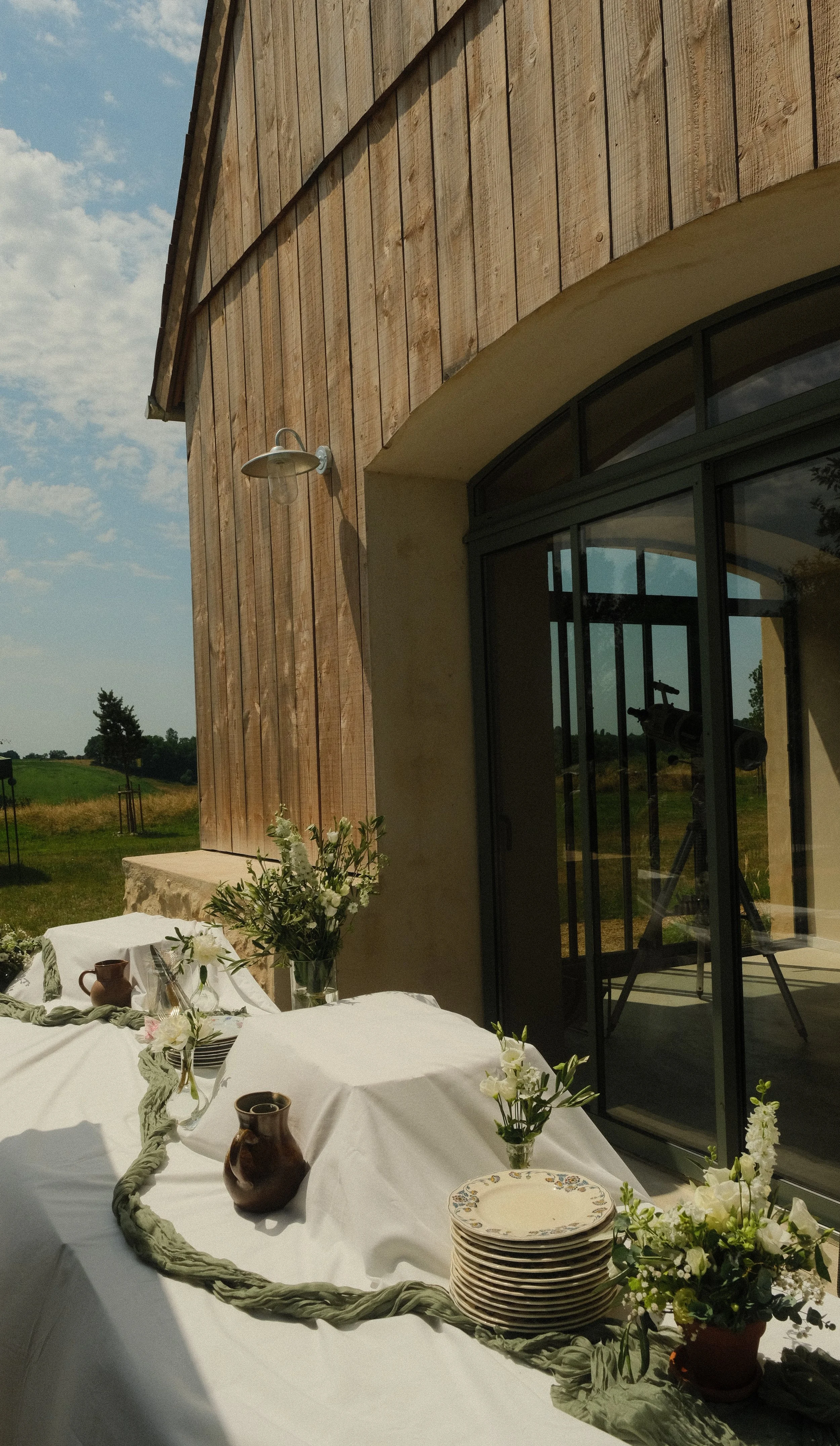 Table dressée avec des assiettes en porcelaine, des vases avec des fleurs blanches, et des tasses en céramique, sous un porche en bois, avec un paysage champêtre en arrière-plan.