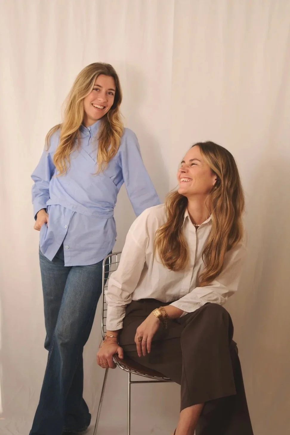 Deux femmes souriantes posent devant un mur blanc, l'une debout et l'autre assise sur une chaise.