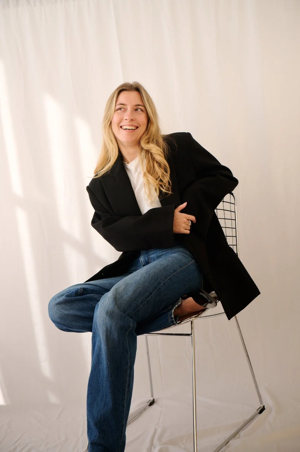 Femme souriante assise sur une chaise en métal, portant un blazer noir et des jeans, dans un fond blanc