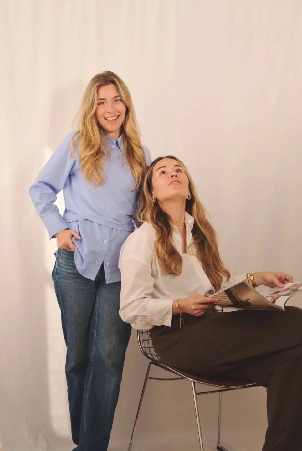 Deux jeunes femmes dans un intérieur clair, l'une debout souriante, l'autre assise, regardant en haut tout en lisant un magazine.