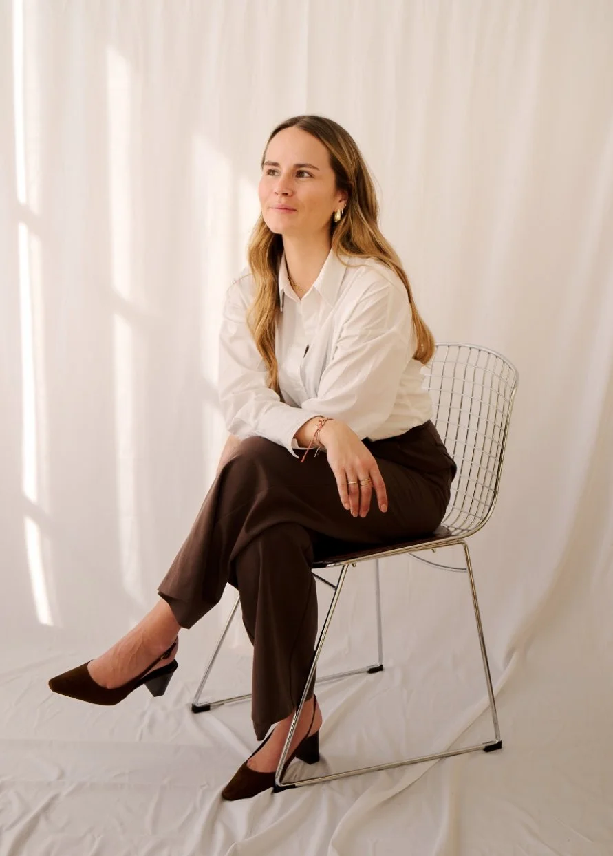 Femme assise sur une chaise en métal, portant une chemise blanche et un pantalon marron, avec un fond blanc