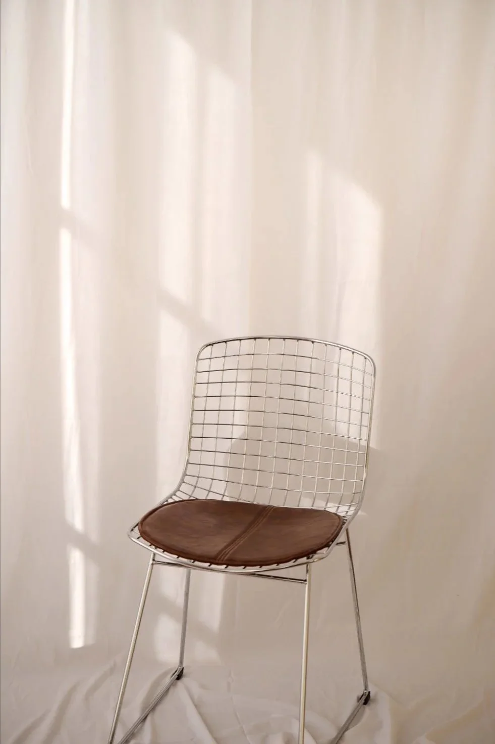 Chaise en fil métallique blanche avec coussin marron au siège, placée contre un mur blanc, avec des reflets de lumière sur le mur.
