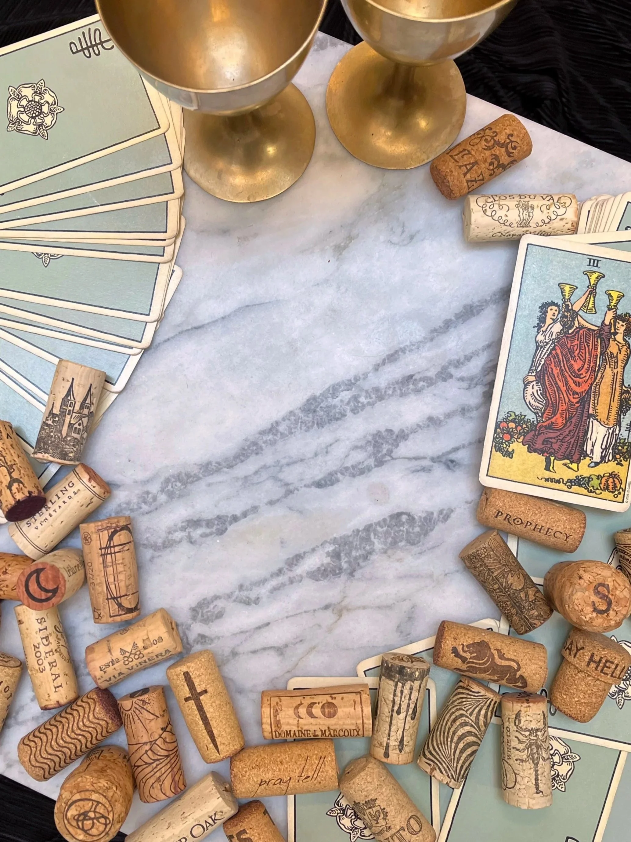 A table with tarot cards, wine corks, and two gold goblets, on a marble surface.