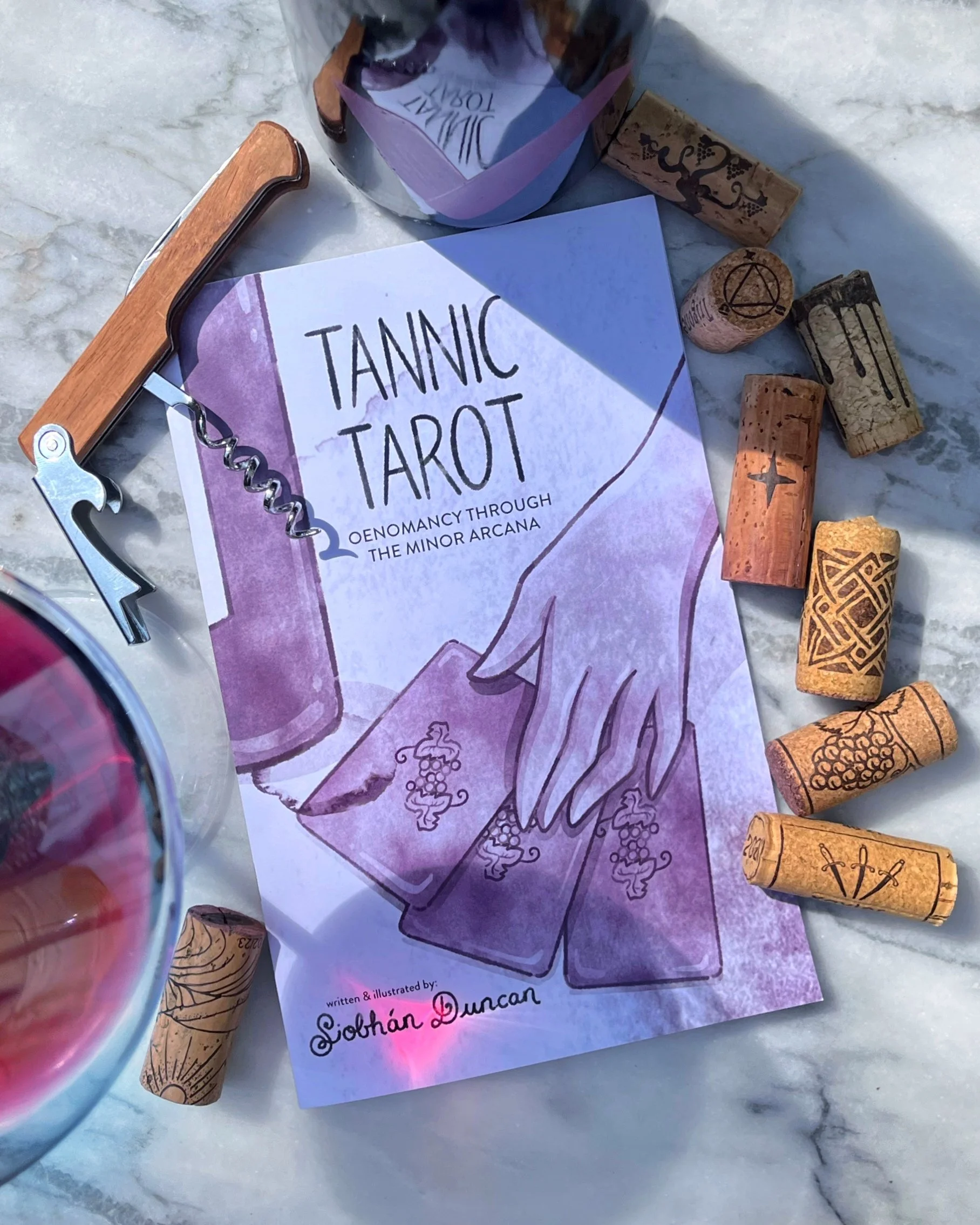A booklet titled "Tannic Tarot" with an illustration of a hand holding tarot cards is surrounded by wine corks and a corkscrew, on a marble surface.