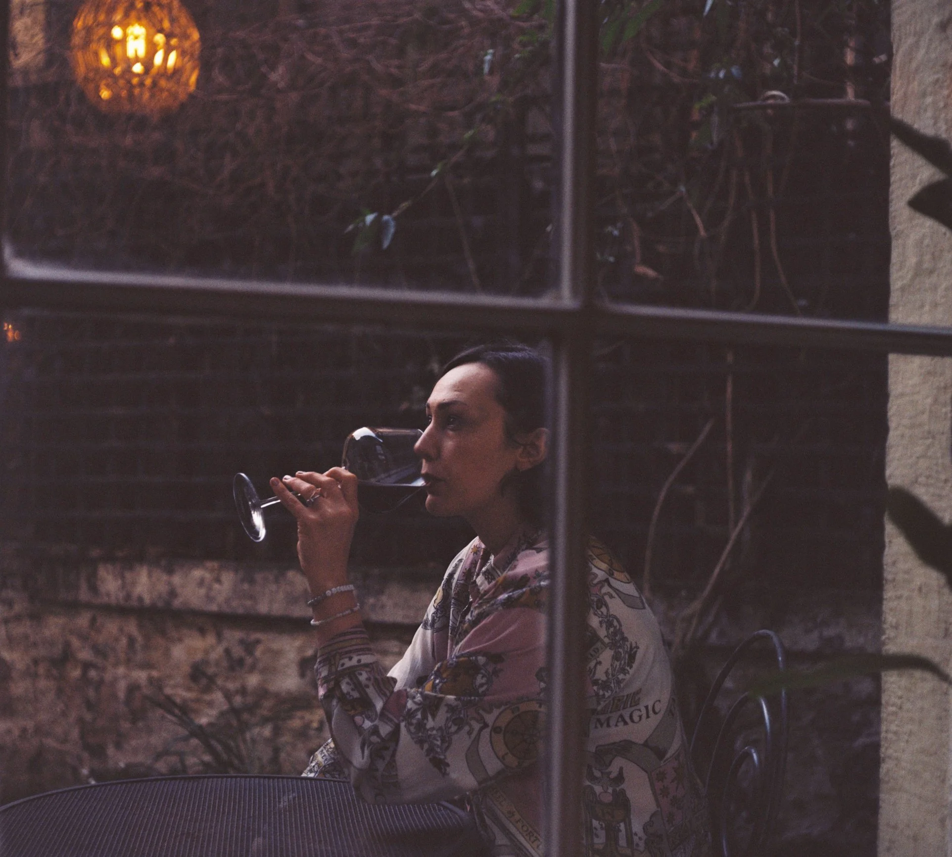 A film photo of a woman sitting at a courtyard table drinking a glass of red wine, viewed through a window pane.
