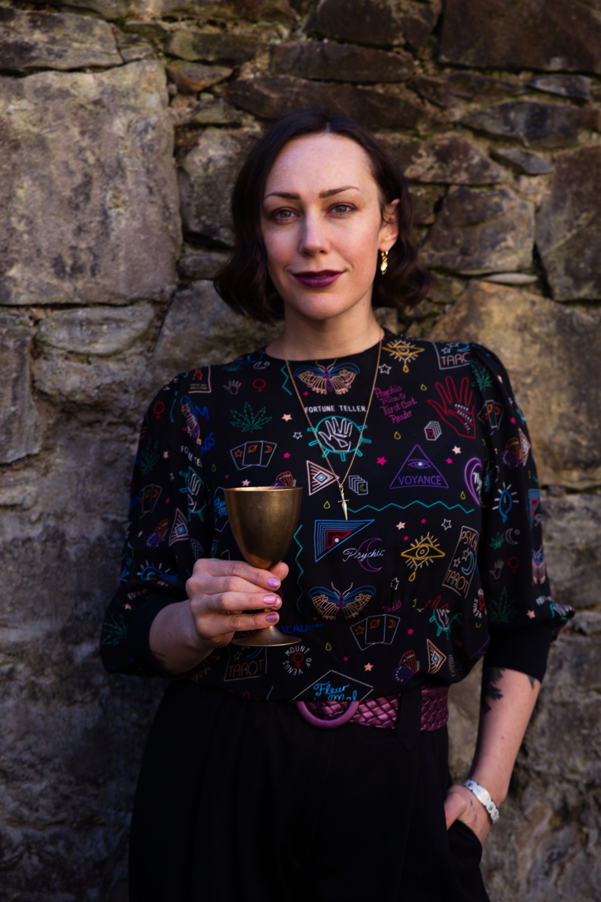 A woman with short dark hair holds a gold goblet, standing in front of a stone wall, wearing a black shirt with colorful mystical and tarot-themed patterns.