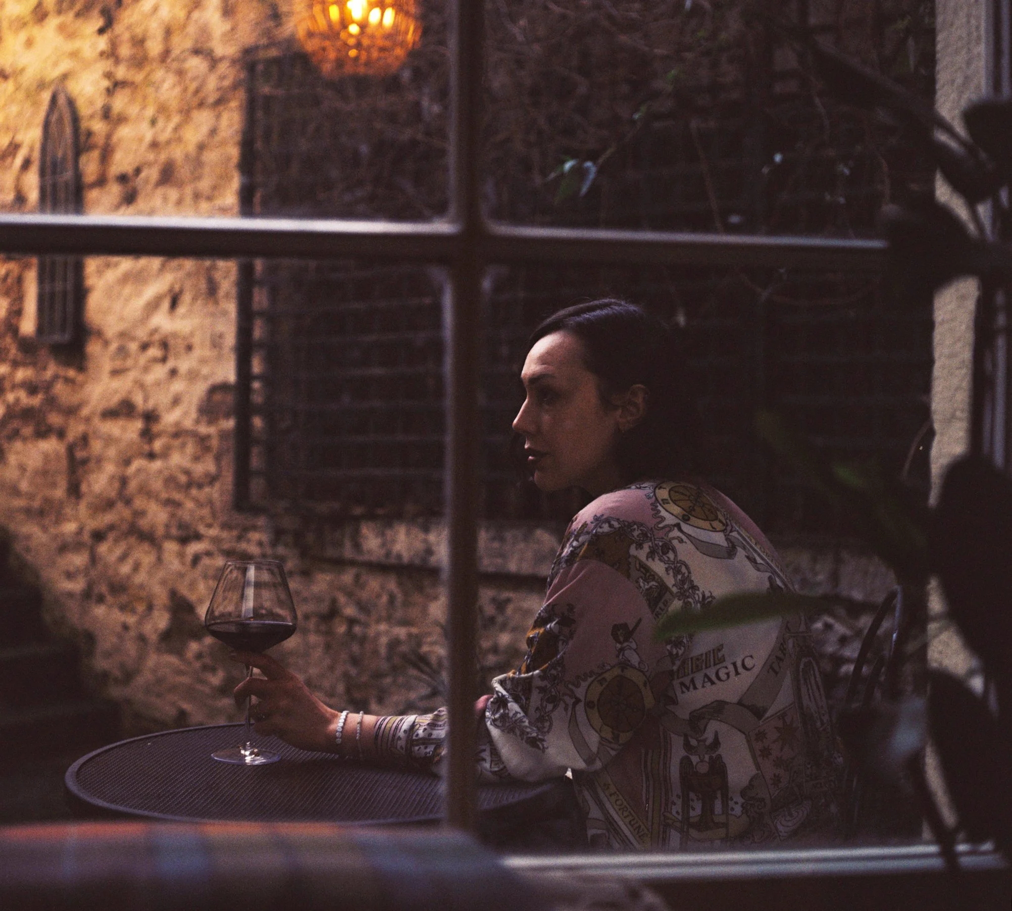 A film photo of a woman sitting at a courtyard table holding a glass of red wine, viewed through a window pane.