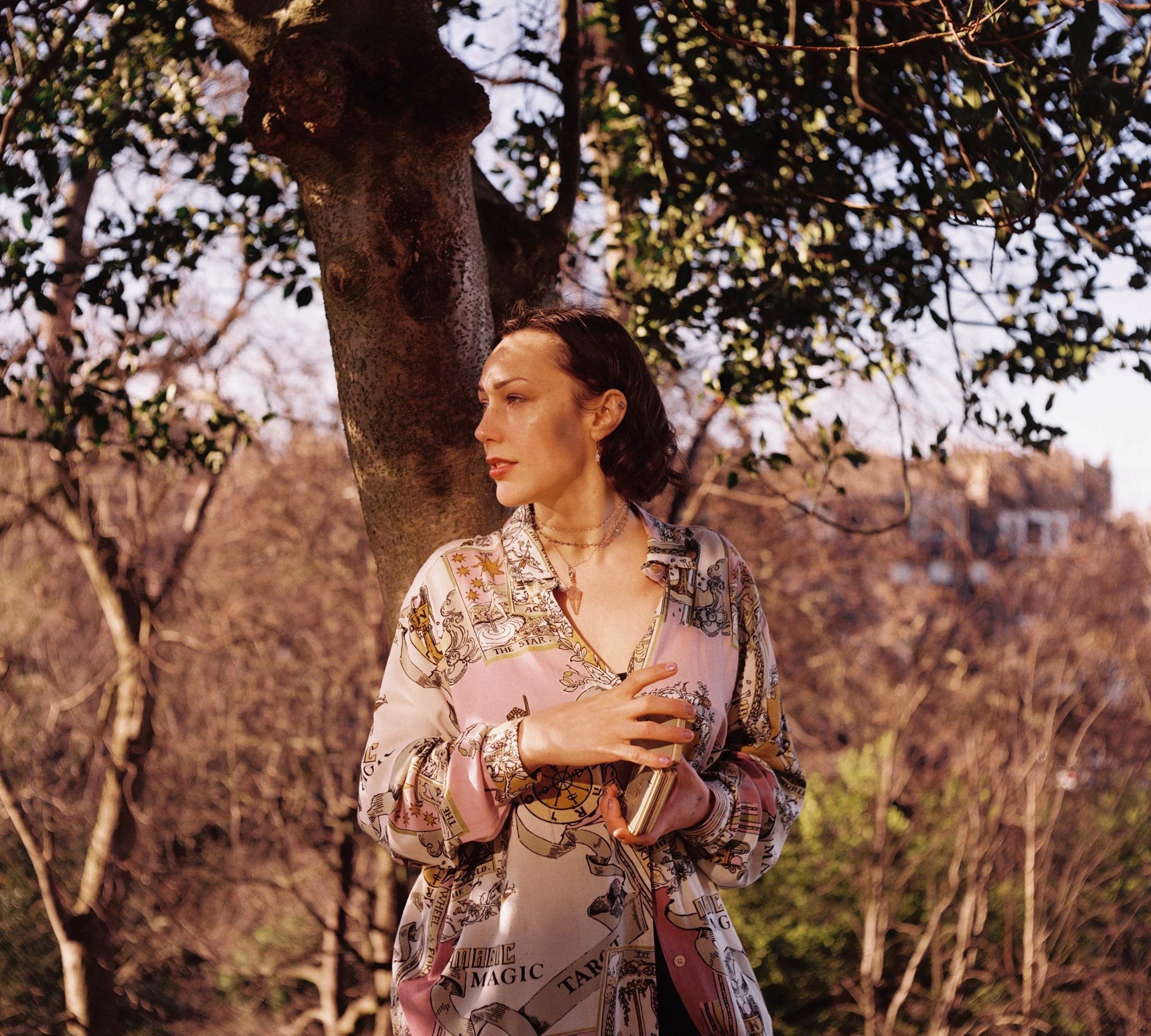 A film photo of a woman with short brown hair wearing a pink tarot-print blouse, holding a deck of tarot cards, standing outdoors near a tree in a garden overlooking Edinburgh in golden afternoon light.