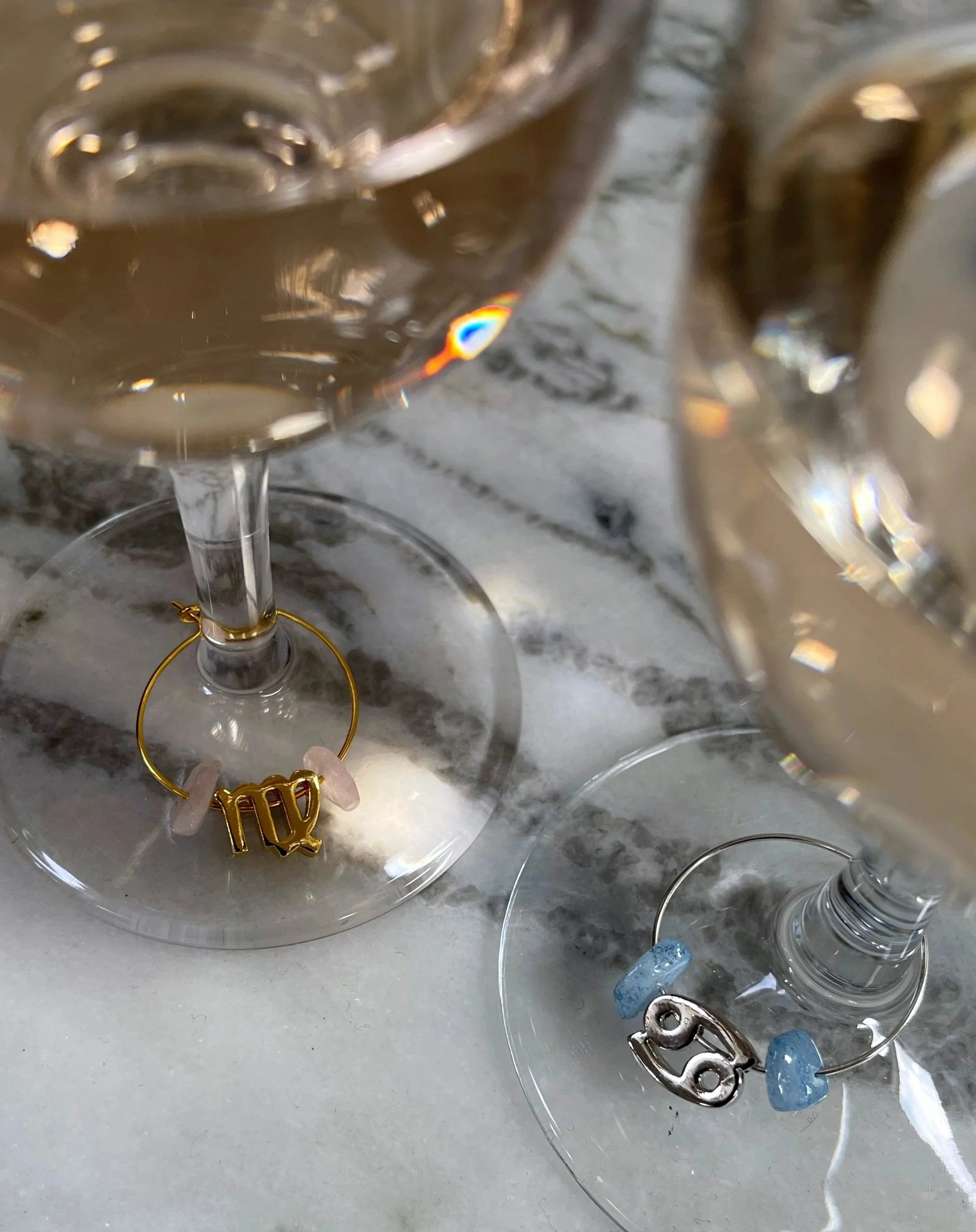 Two wine glasses filled with rosé wine have decorative rings around the stems, one with a gold Virgo glyph and rose quartz stones, the other with silver Cancer glyph and aquamarine stones, placed on a marble surface.
