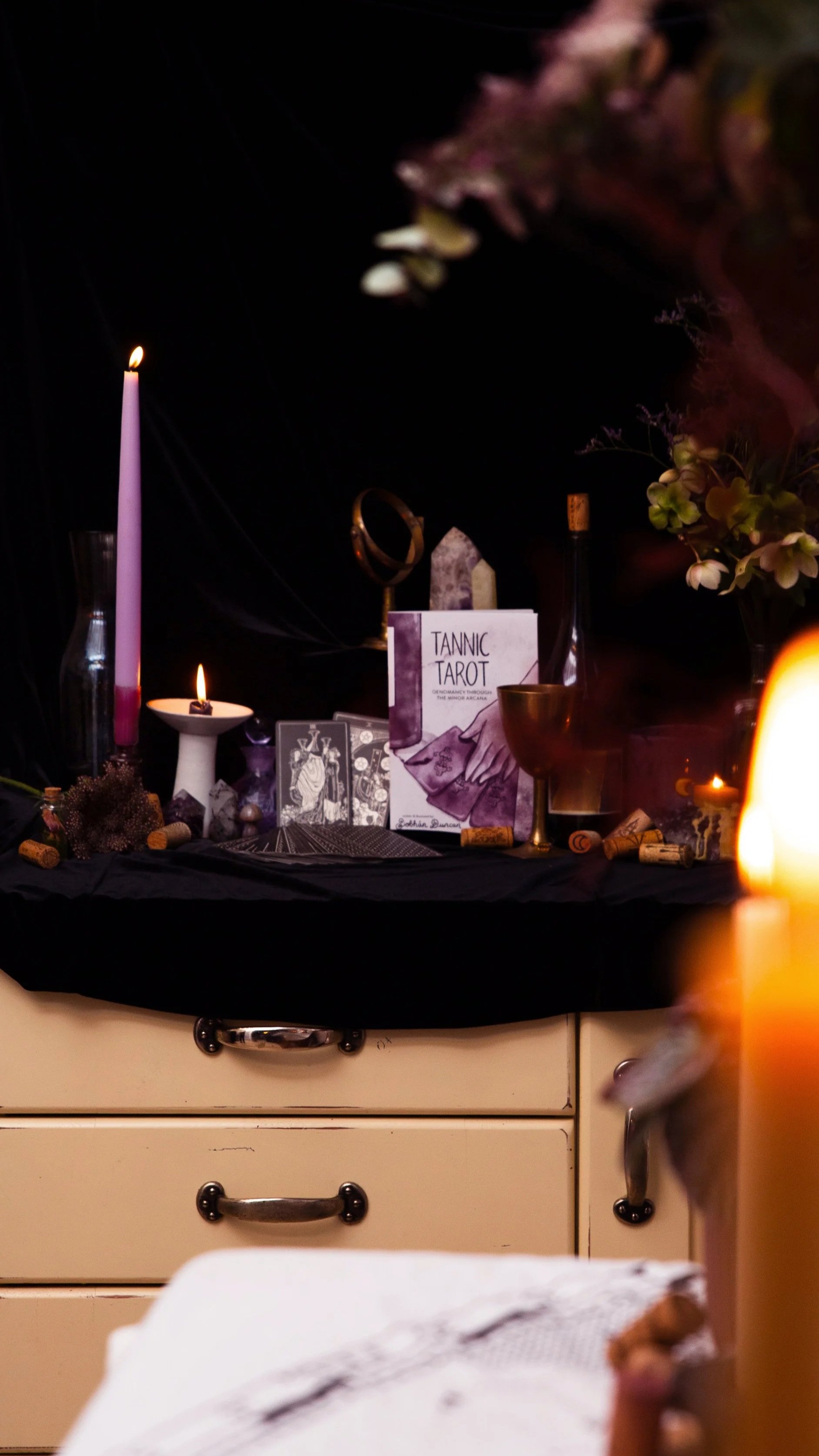 A small altar with candles, tarot cards, a mirror, books, and flowers on a black cloth draped over a table. The scene is dimly lit with a dark background.