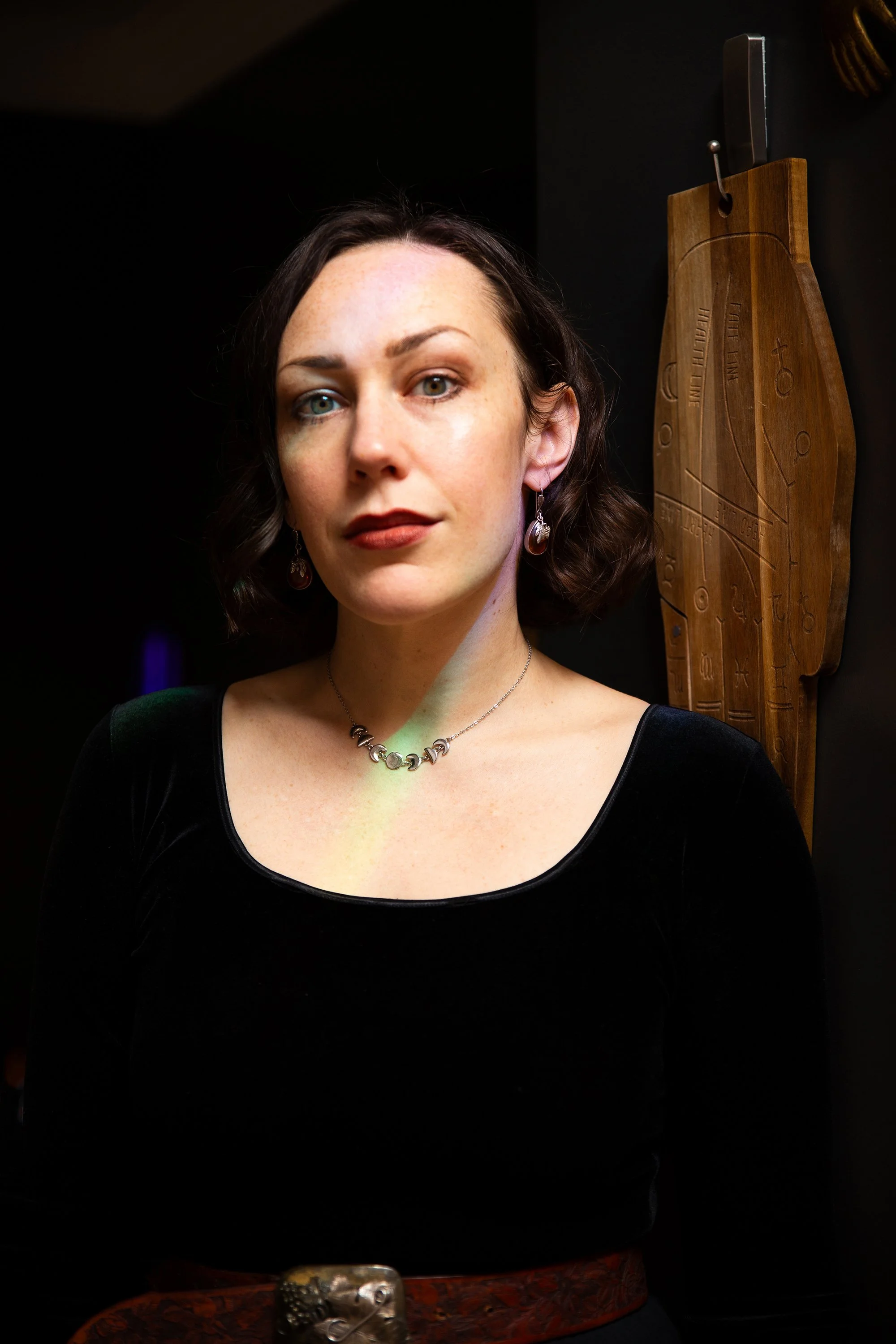 A woman with short brown hair and green eyes in a black dress, standing indoors against a dark background with a wooden palmistry hand mounted on the wall behind her.