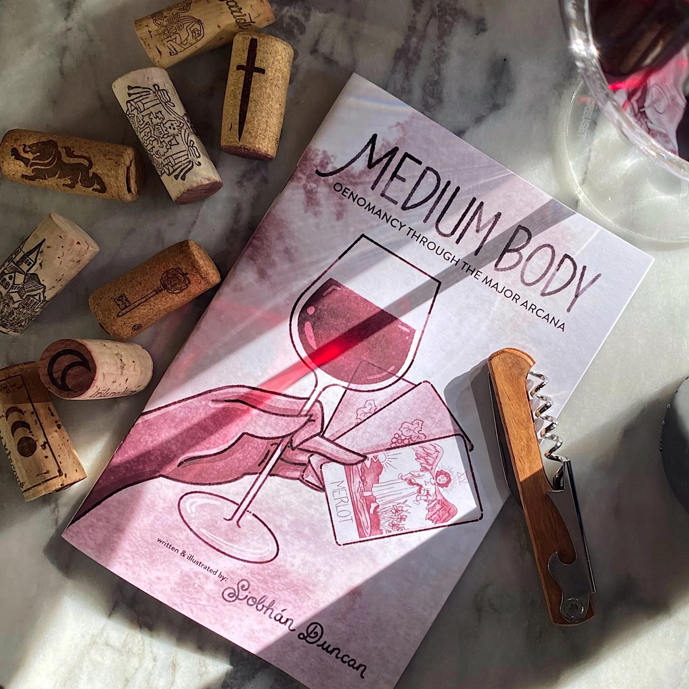 A booklet titled "Medium Body" with an illustration of a hand holding a glass of red wine and a tarot card. Surrounding corks and a corkscrew, on a marble surface.