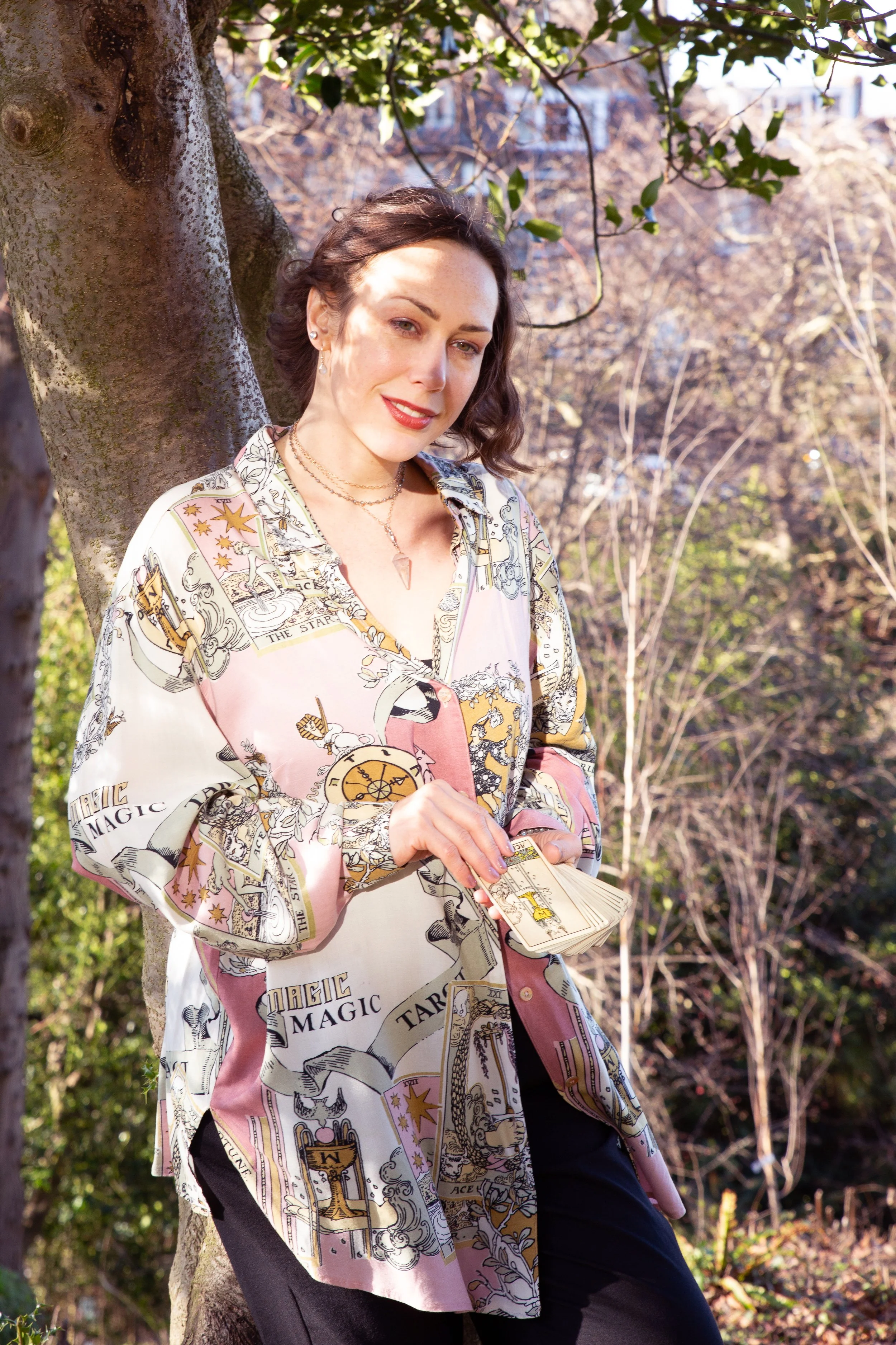 A woman with short brown hair dressed in a pink tarot-themed shirt, stands outdoors leaning against a tree, holding a few tarot cards in her hand, with a background of trees and sunlight.