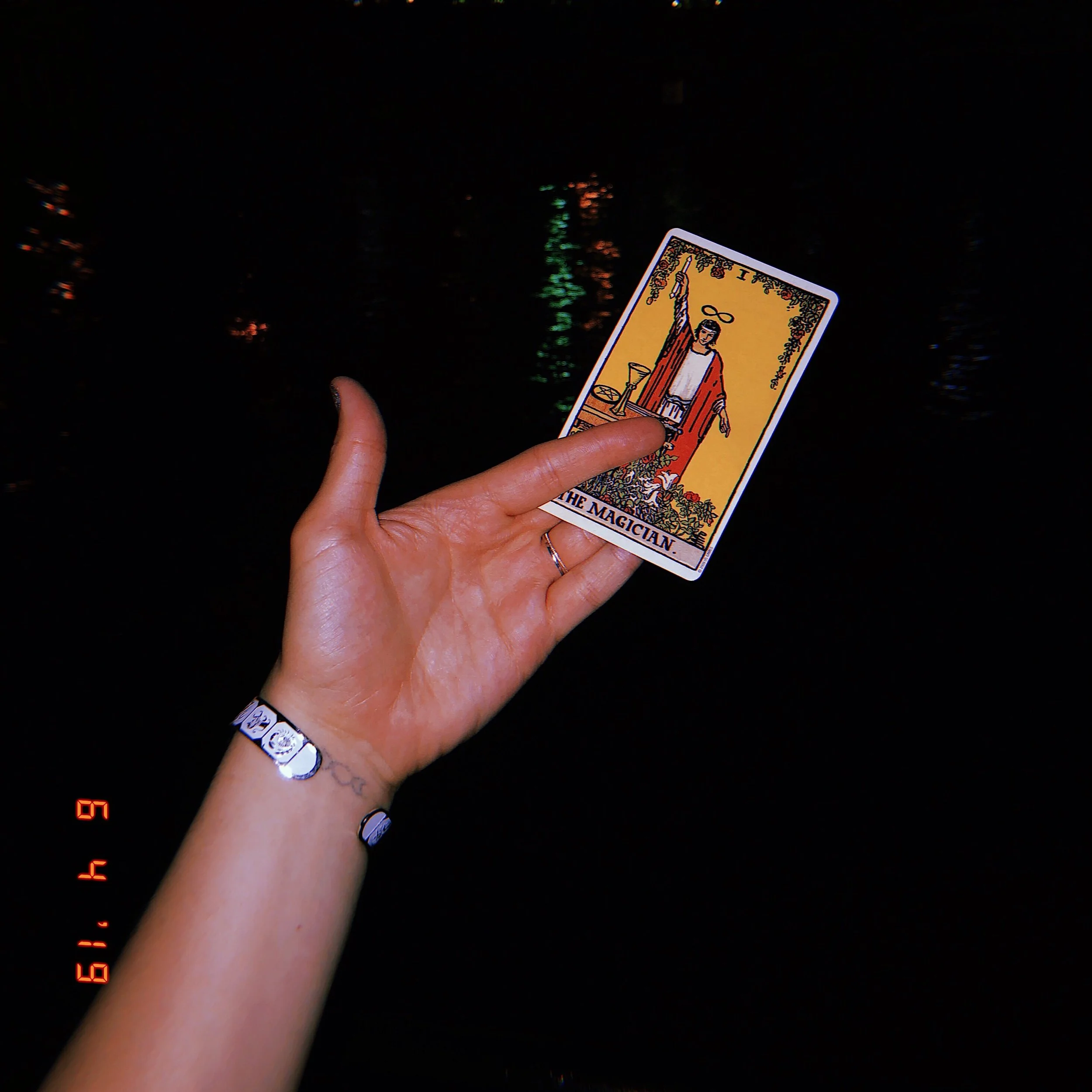 A person holds a tarot card labeled 'The Magician' with a dark, water-like background with reflections.