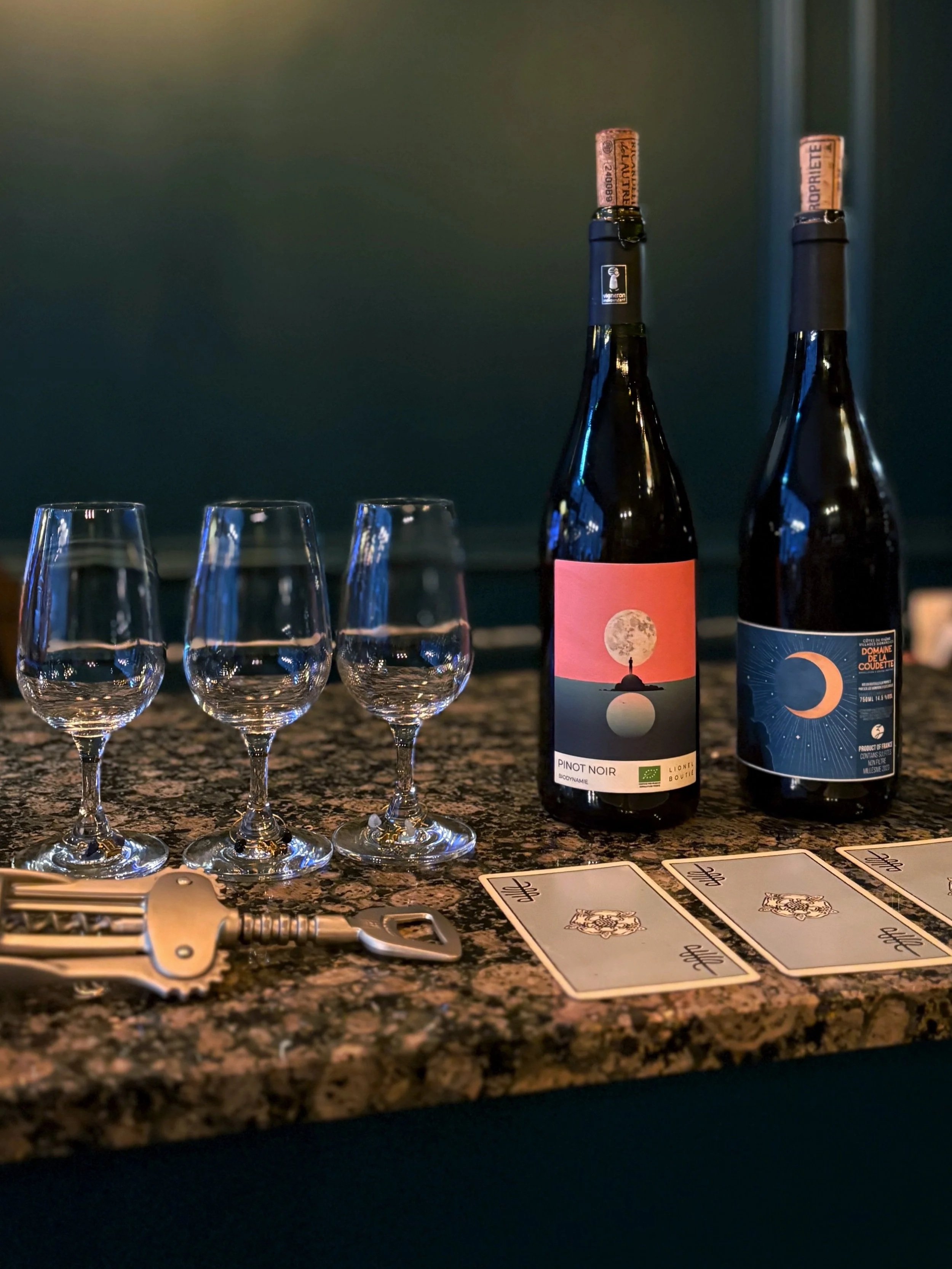 Two bottles of wine, three empty wine glasses, and three tarot cards on a shop countertop with a dark background.
