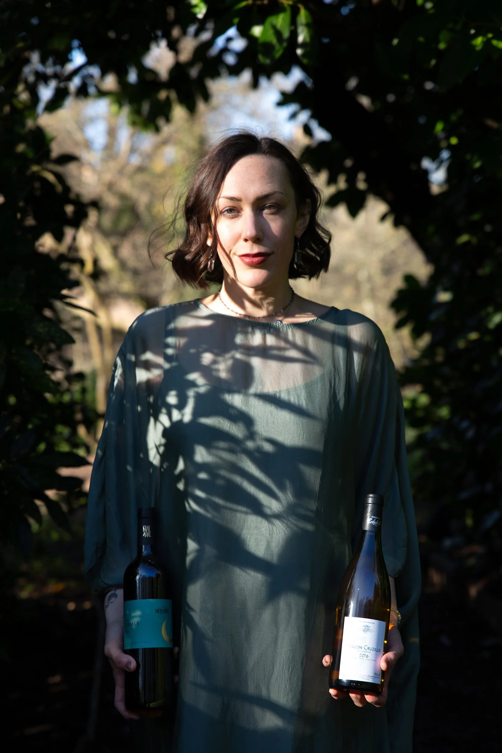 A woman stands in a garden in sunlight and shadow, holding a bottle of wine in each hand. She has short brown hair, wears a sage green tunic, and has a subtle smile.