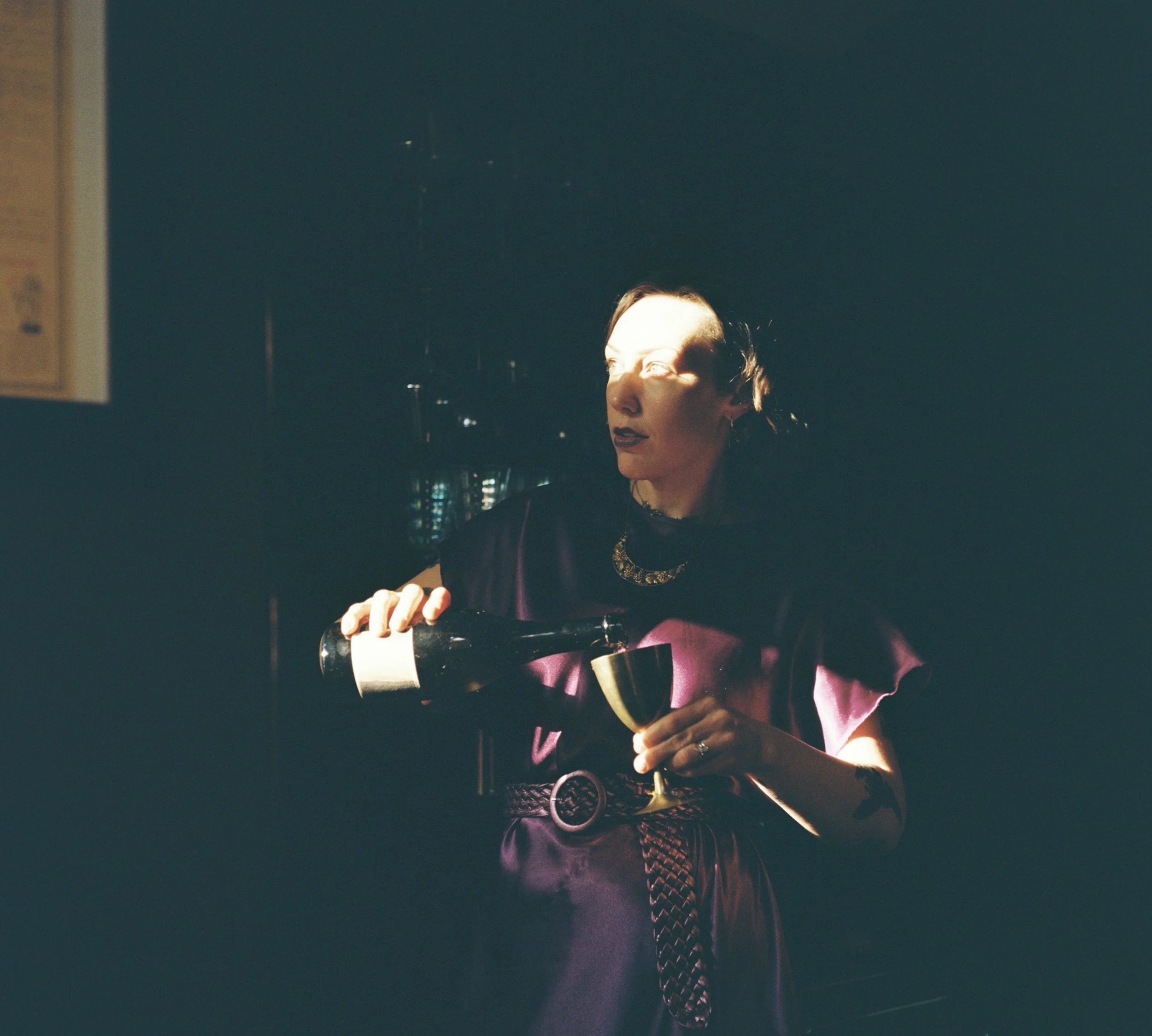 A woman with short hair in a dark purple dress pours wine into a glass, illuminated by a beam of light, with a dark background.