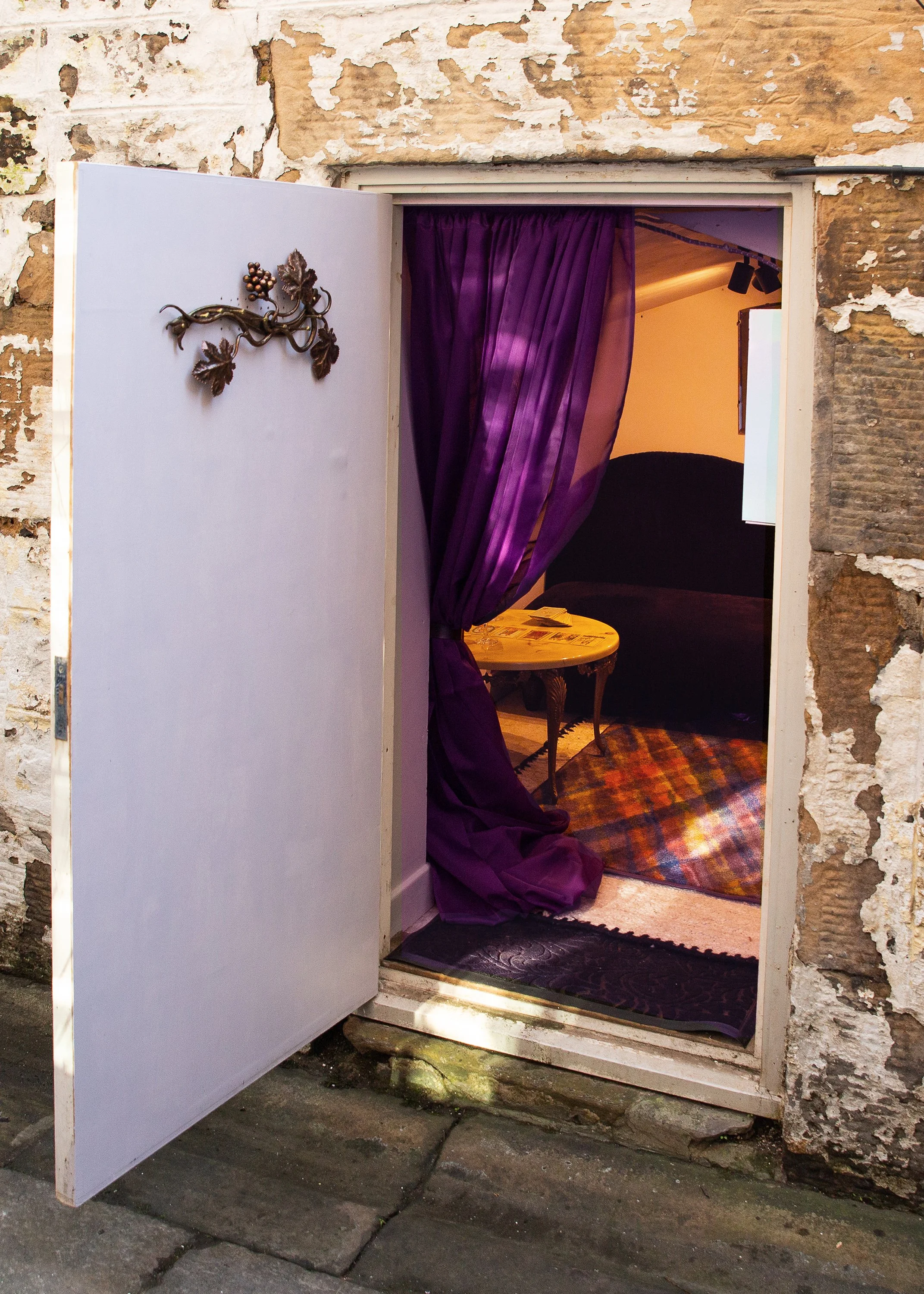 A small door with a purple curtain slightly open, revealing a cozy room with dark furniture, a pouf, a patterned rug, and a small table, against an exterior wall with peeling paint.
