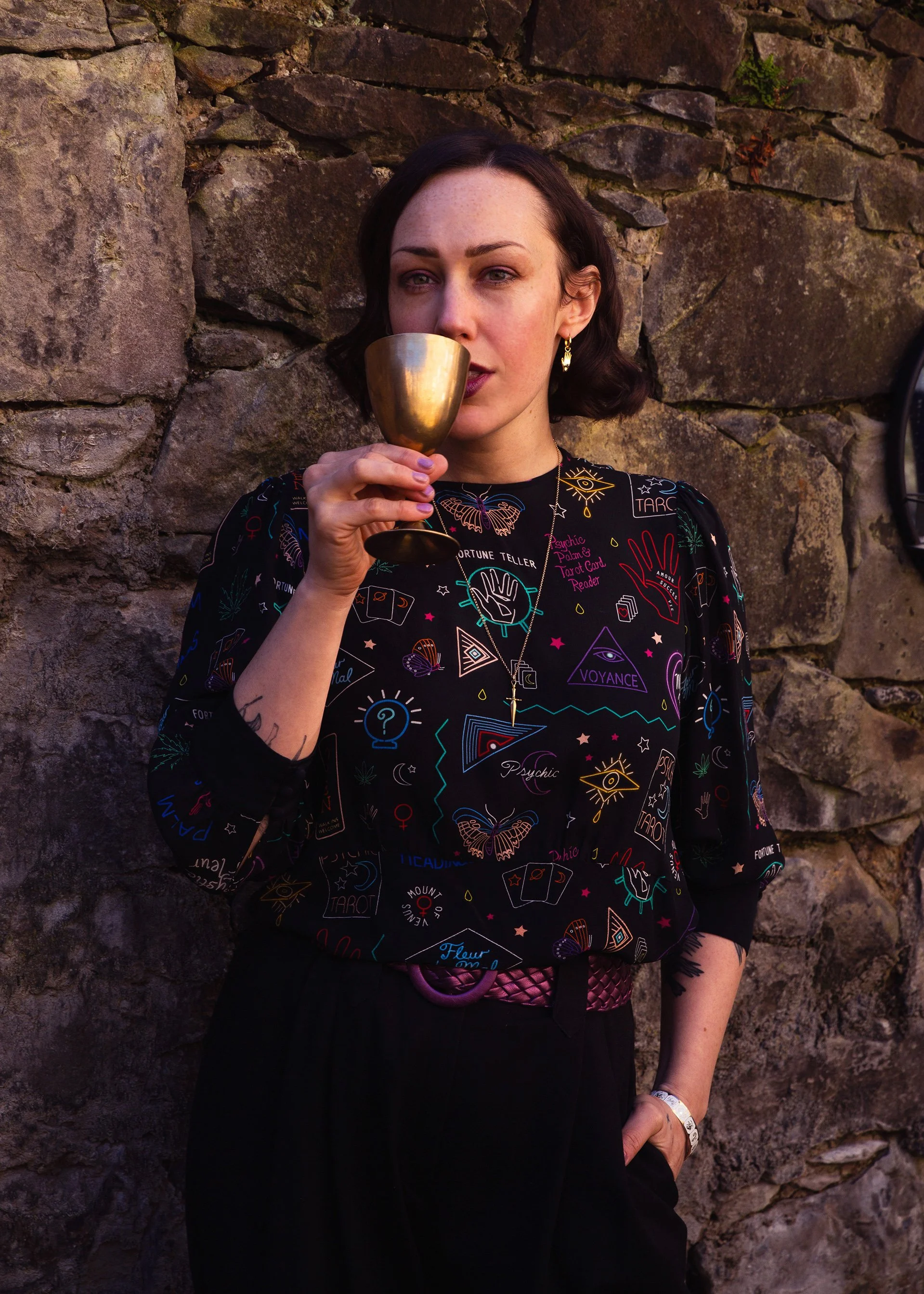 A woman with short brown hair holds a gold cup close to her lips, standing against a stone wall. She wears a black top decorated with colorful tarot and mystical symbols.