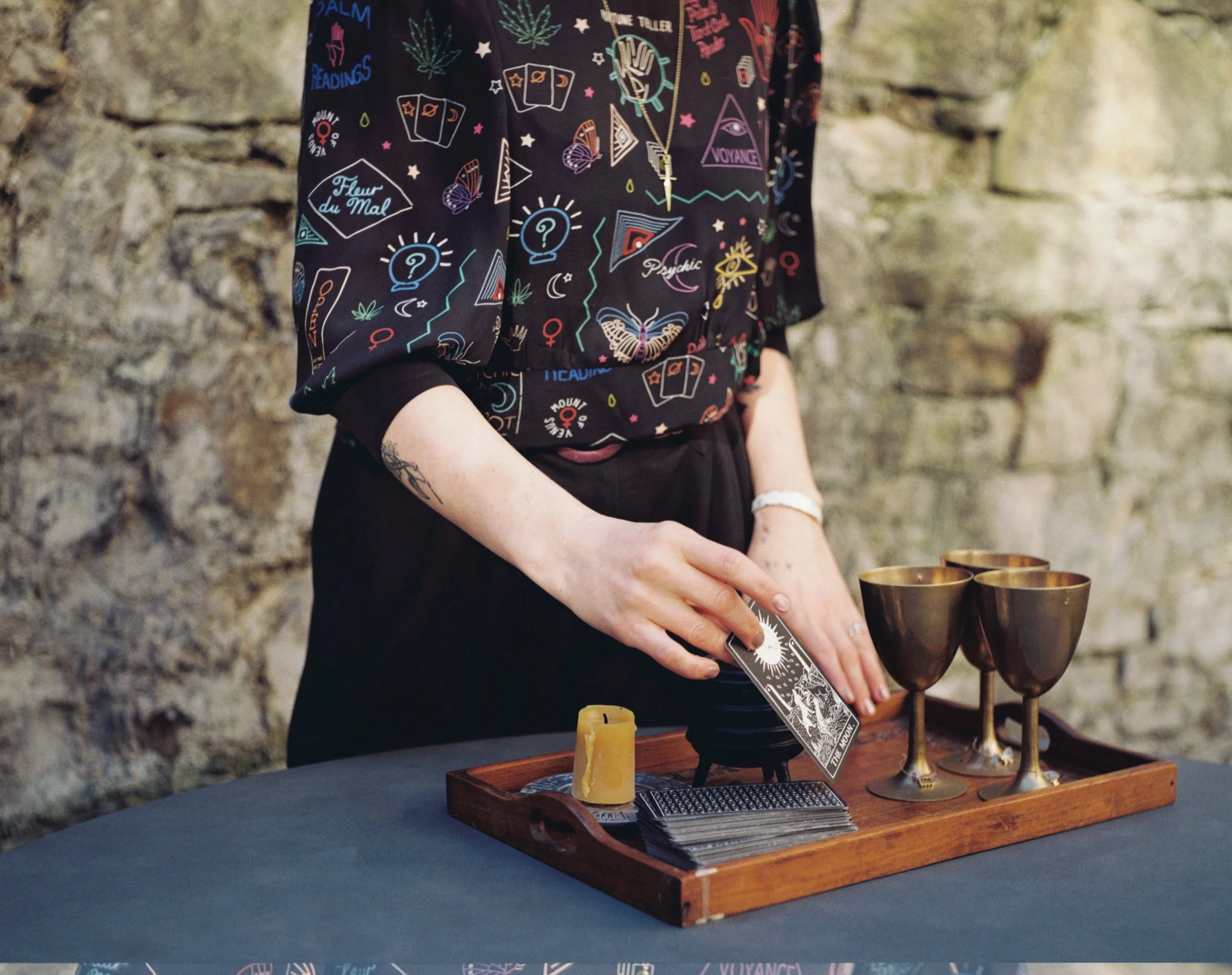 The torso of a woman wear a tarot print shirt, standing at a table with a wooden tray holding three gold goblets, tarot cards, and a beeswax candle, with a stone wall background.