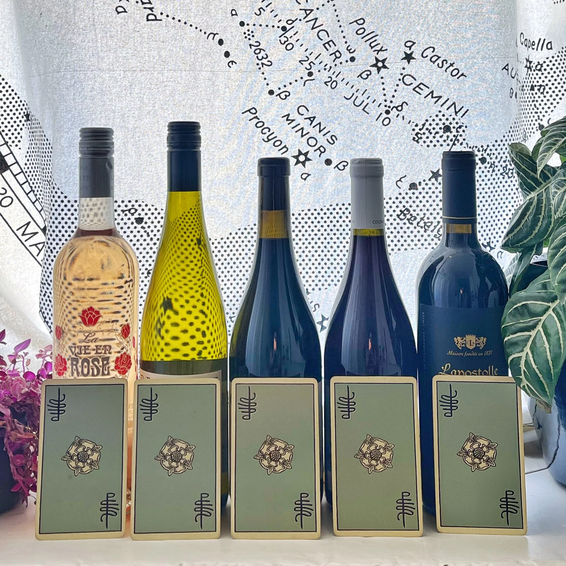 Five bottles of wine are displayed in a row, with tarot cards placed in front of each bottle against a backdrop of a star map.