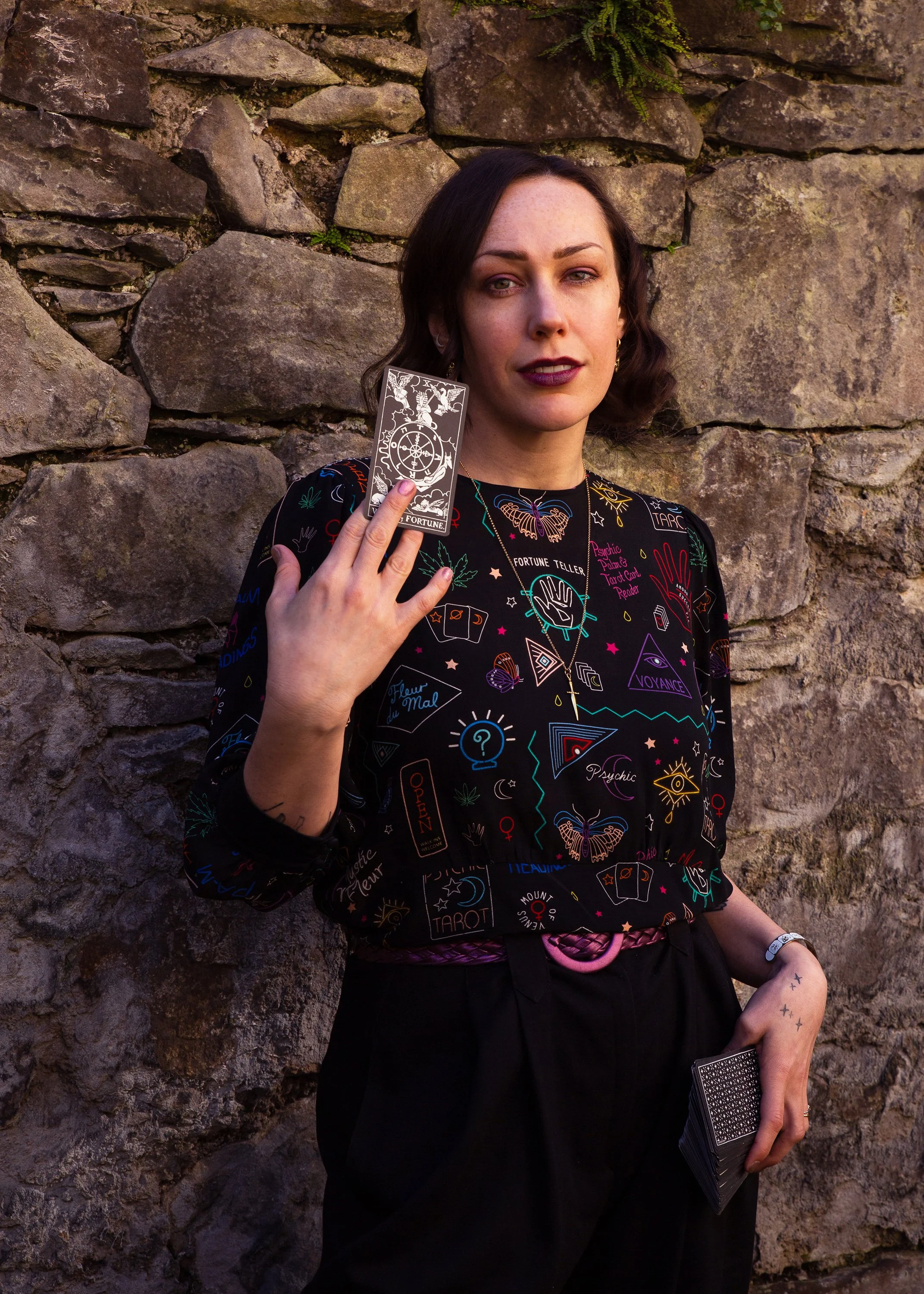 A woman with dark hair, dark lipstick, and tattoos on her fingers, standing against a stone wall, holding tarot cards, with a mystical and occult-themed outfit covered in symbols and drawings.