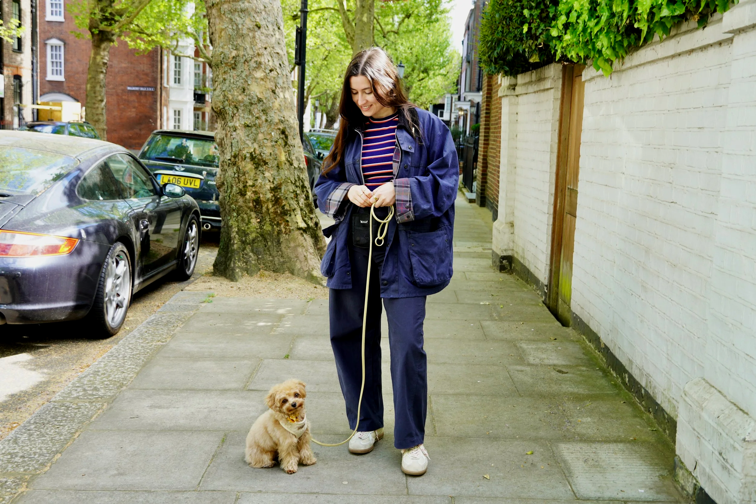 A young woman walking her small dog on a leash on a city sidewalk, surrounded by parked cars and green trees, with brick and painted walls in the background.
