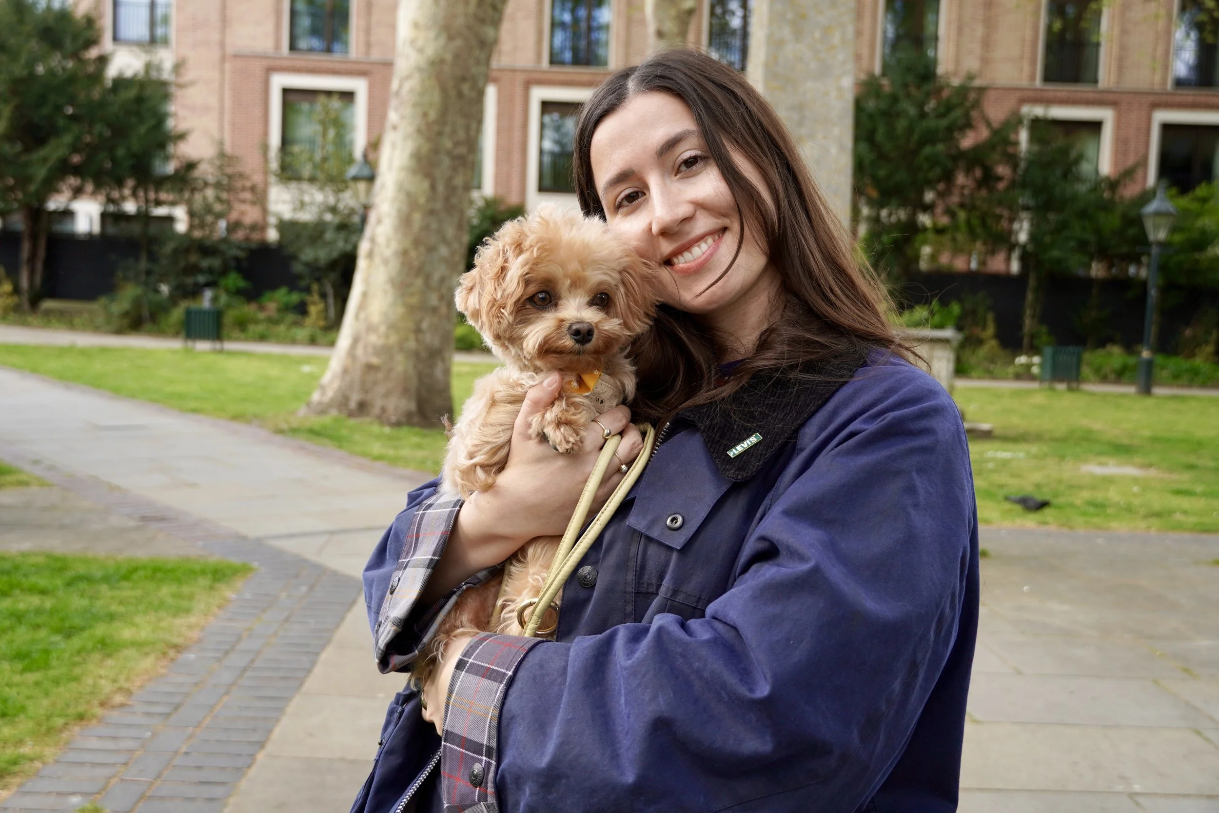A woman smiling and holding a small tan-colored dog outdoors in a park with trees and residential buildings in the background.