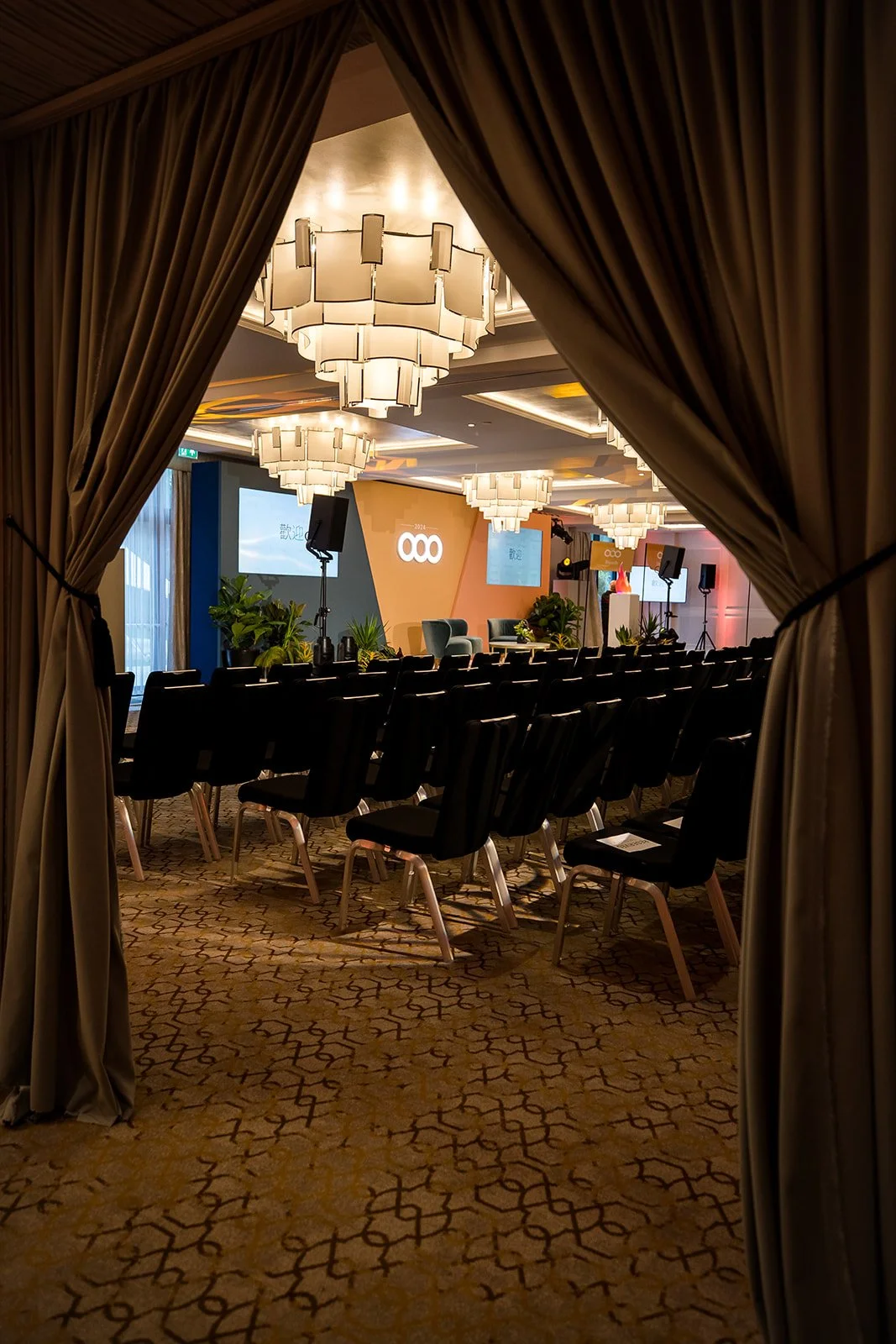 View of an event stage seen through partially opened curtains, with plush chairs, plants, large chandeliers, a projection screen, and a logo on a backdrop in a conference room setting.