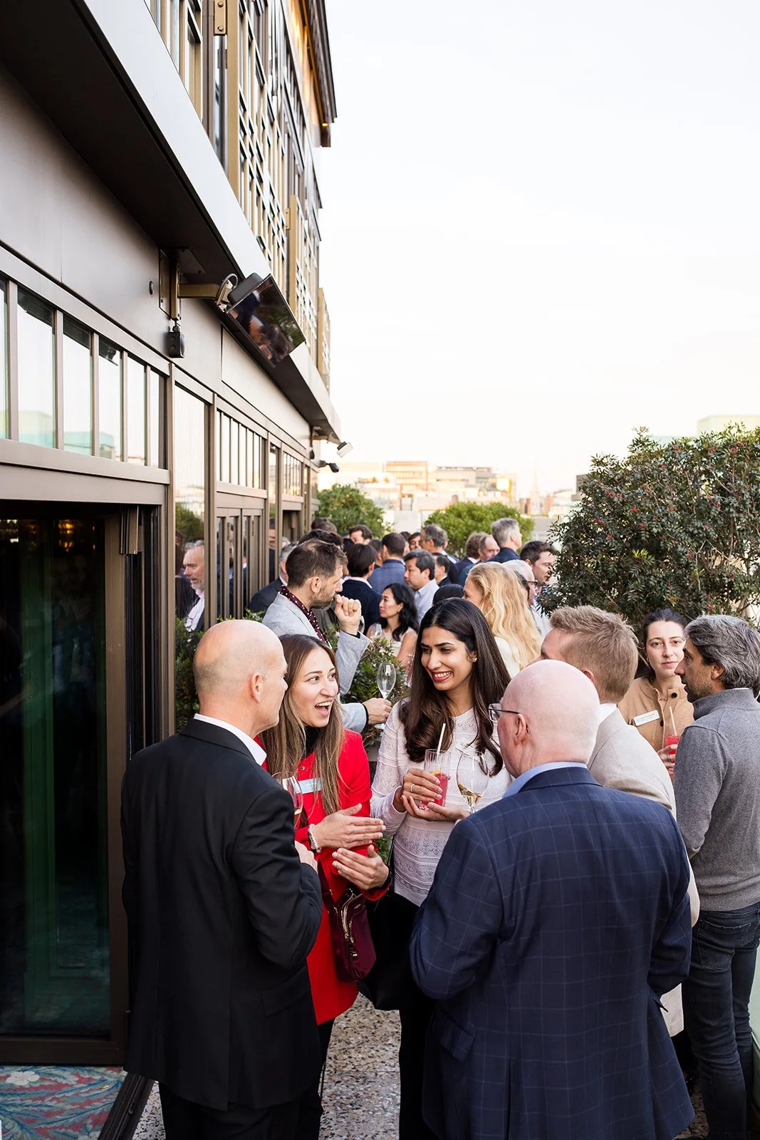 A group of people gathered outside on a balcony, socializing and holding drinks, at a rooftop event during the evening.