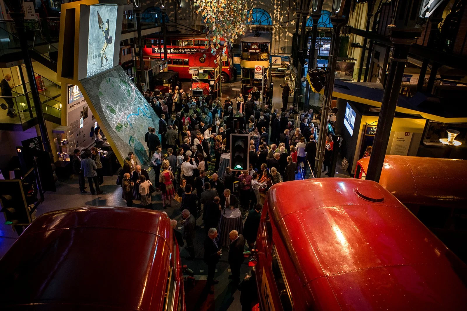 A crowded indoor event with many people socializing and exploring displays, including vintage buses, a large digital map, and various informational booths, inside a multi-level building.