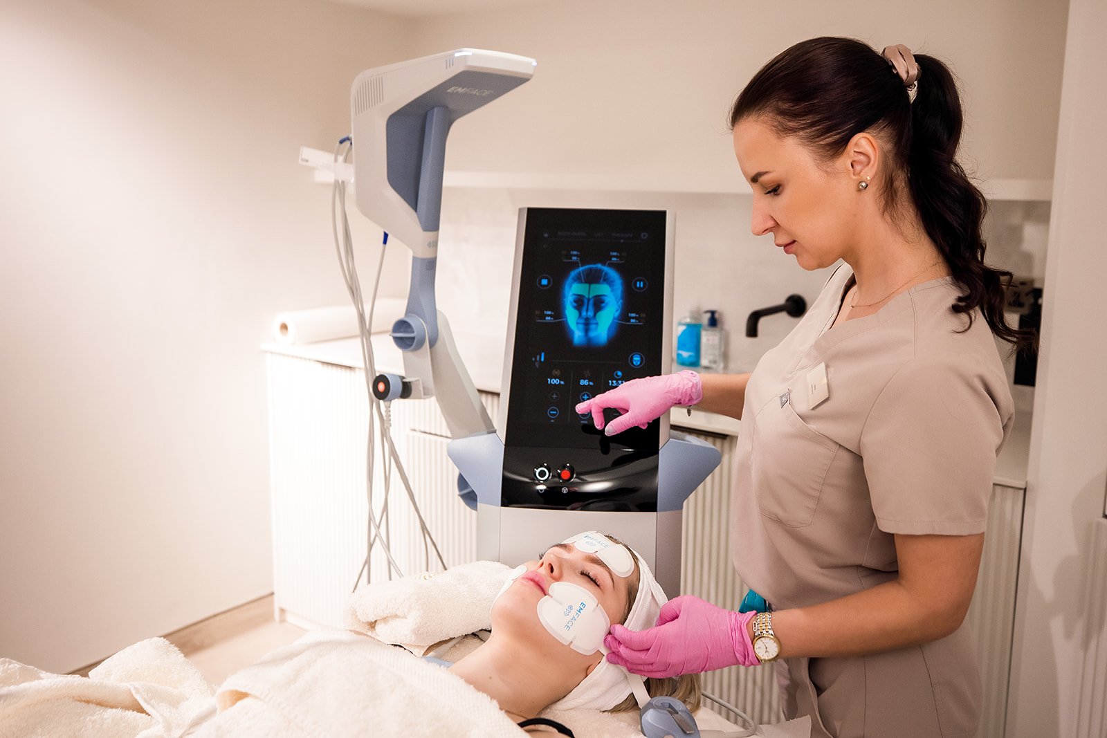 A woman is lying on a bed with medical eye patches, while a healthcare professional is adjusting a high-tech medical device connected to her face. The device displays a monitoring screen with a digital face and control options, indicating a medical o