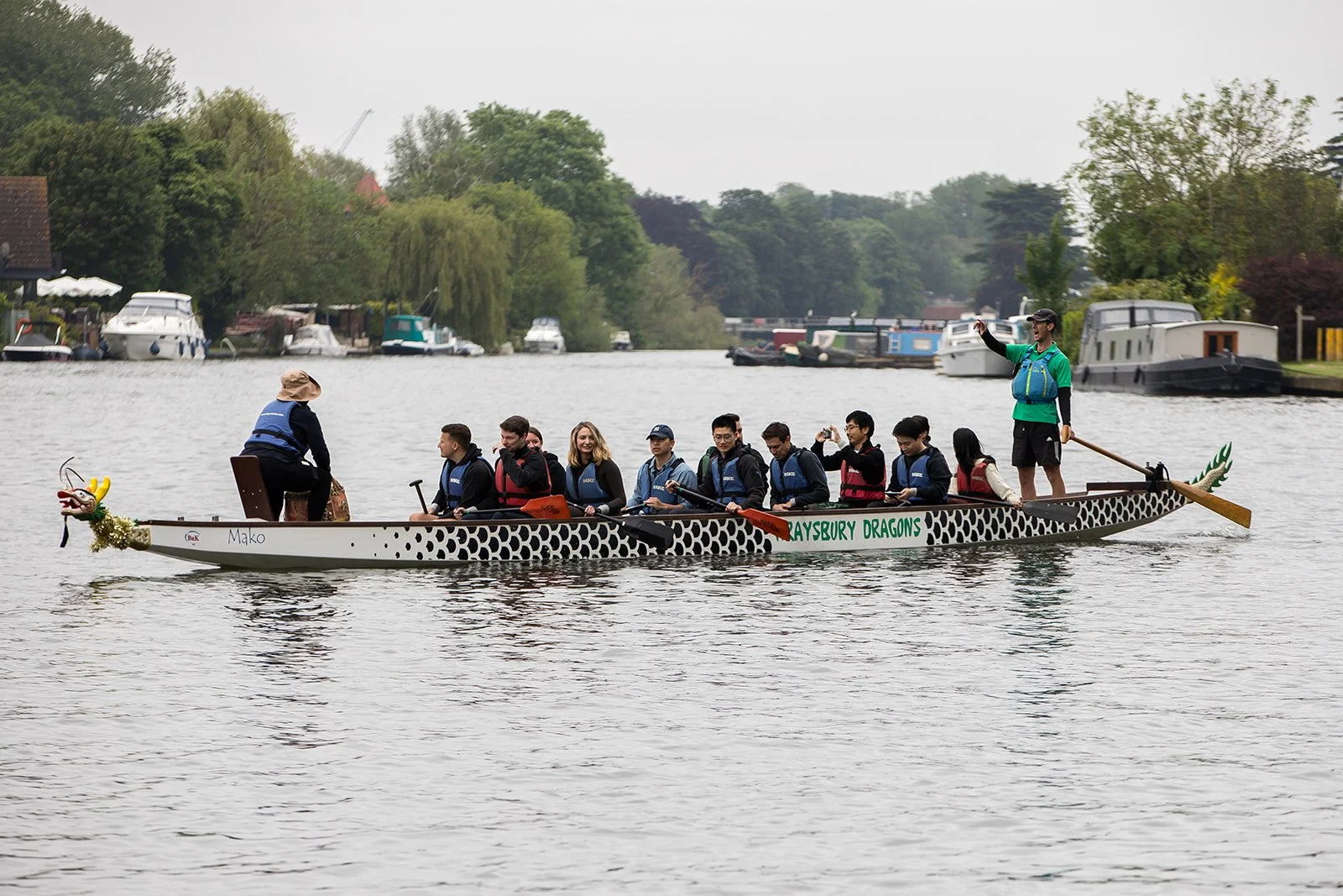 Group of people on a dragon boat racing on a lake, with a person at the front and another at the back paddling, and a woman sitting at the front, near a small decorative dragon head. The boat has 