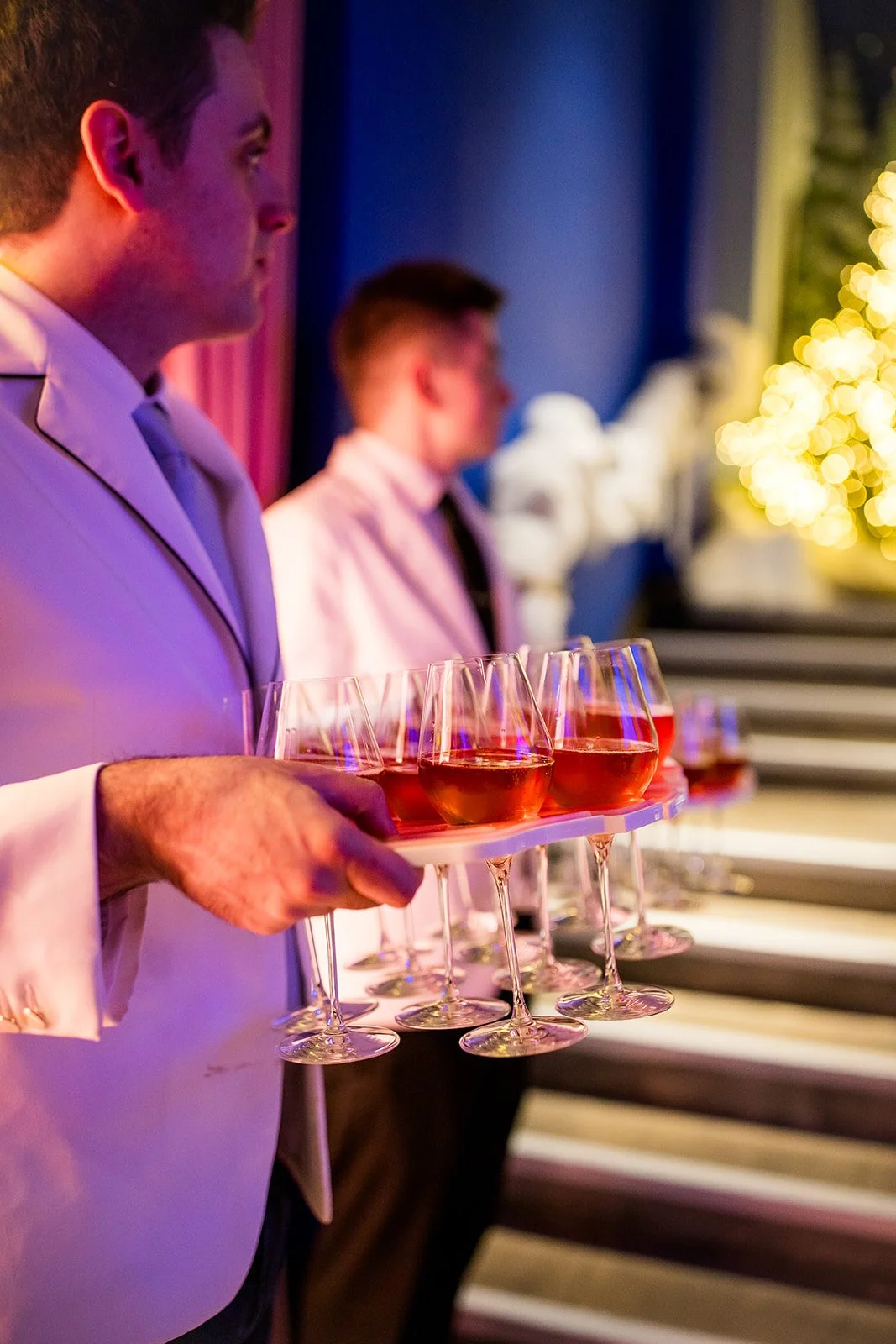 A waiter in a white suit holding a tray with several glasses of pink-colored wine at a party or formal event.
