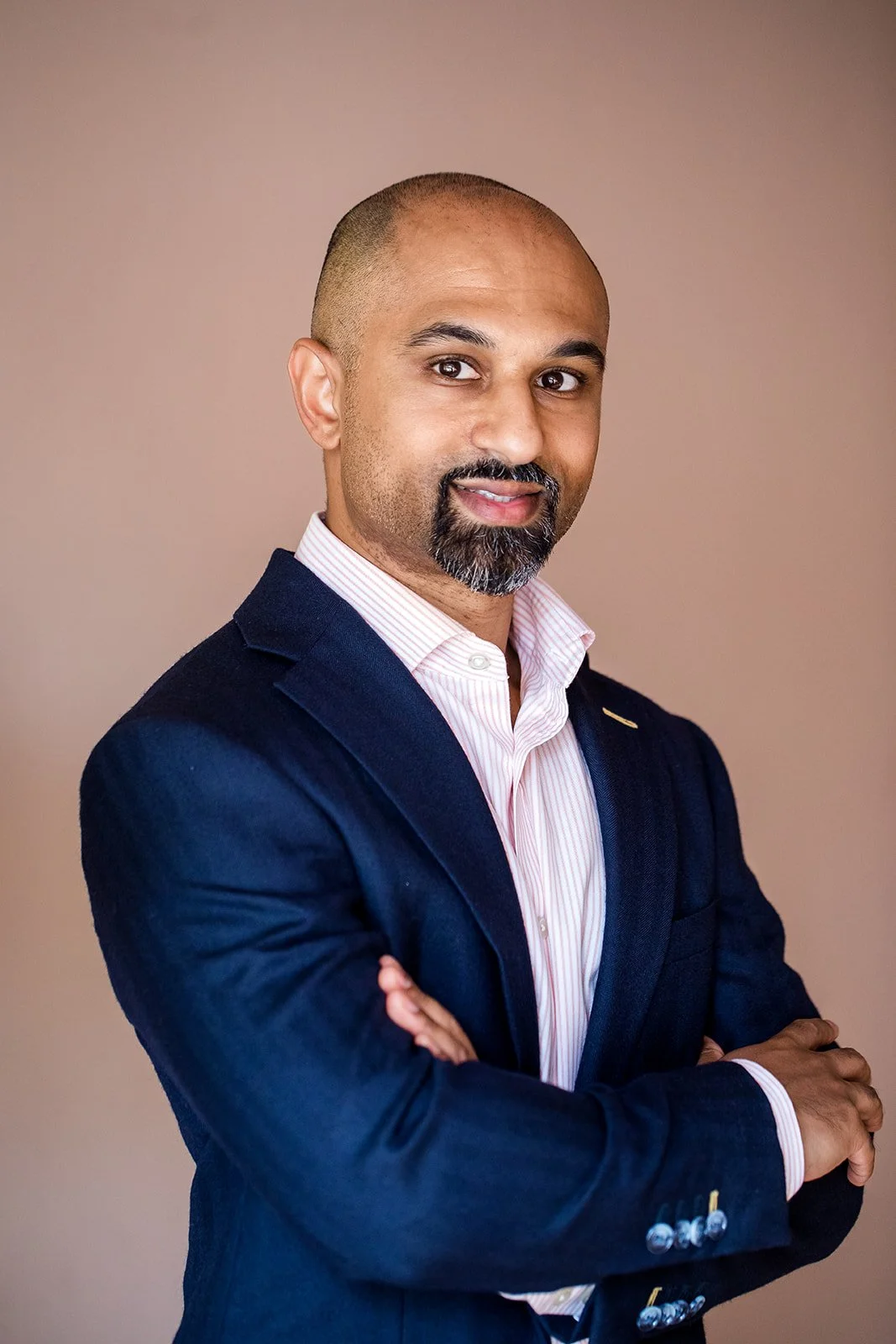 Headshot of a man with a bald head and beard, wearing a dark blue blazer and a light pink striped shirt, standing with arms crossed and smiling at the camera against a plain background.
