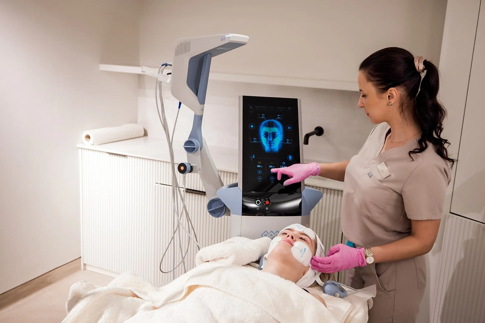 A woman lying on a treatment bed connected to a medical device, receiving a facial scan from a healthcare professional. The professional is using a touchscreen console with a blue facial graphic on display.