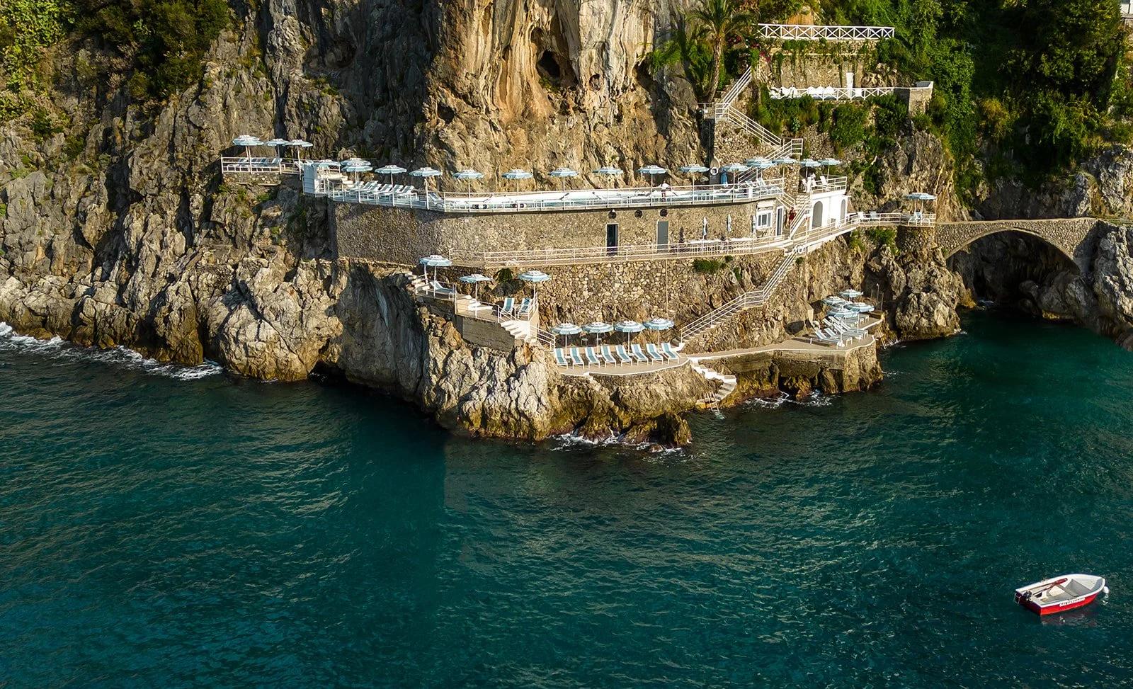 A scenic view of a rocky coastline with multiple levels of white railings, stairs, and terraces featuring blue and white lounge chairs and umbrellas overlooking the ocean. A small boat is floating nearby in the water, and greenery is present on the c