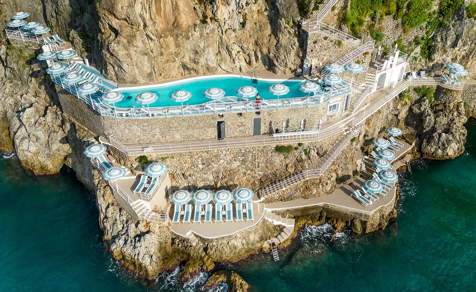 Aerial view of a cliffside swimming pool with multiple lounge chairs and umbrellas, overlooking the ocean.