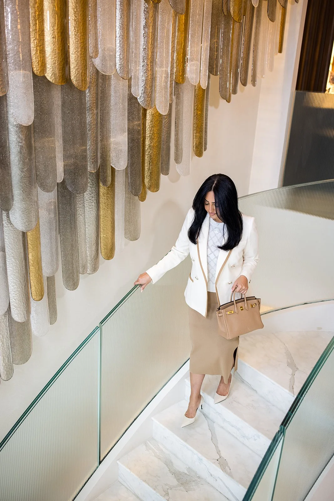 A woman with black hair wearing a white blazer, beige skirt, and high heels walking up a staircase with a glass railing, holding a beige handbag, with a decorative wall hanging of metallic elongated oval shapes in gold, silver, and bronze tones behin