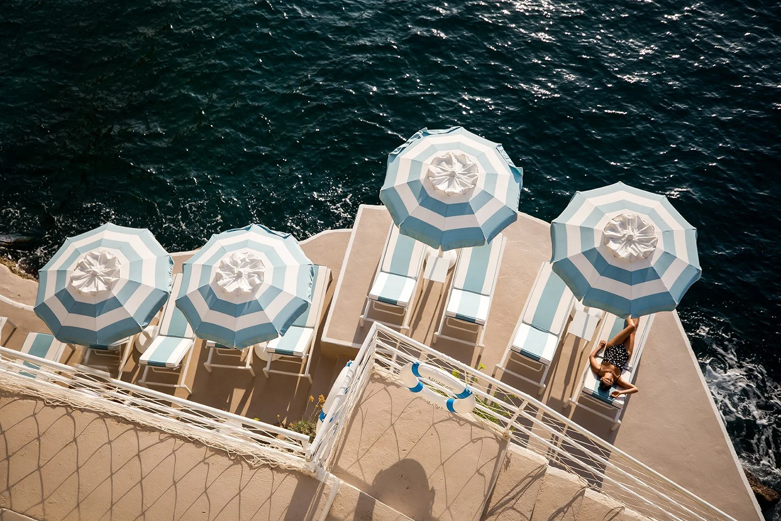 A woman lying on a lounge chair on a balcony overlooking the ocean with four blue and white striped umbrellas and lounge chairs.