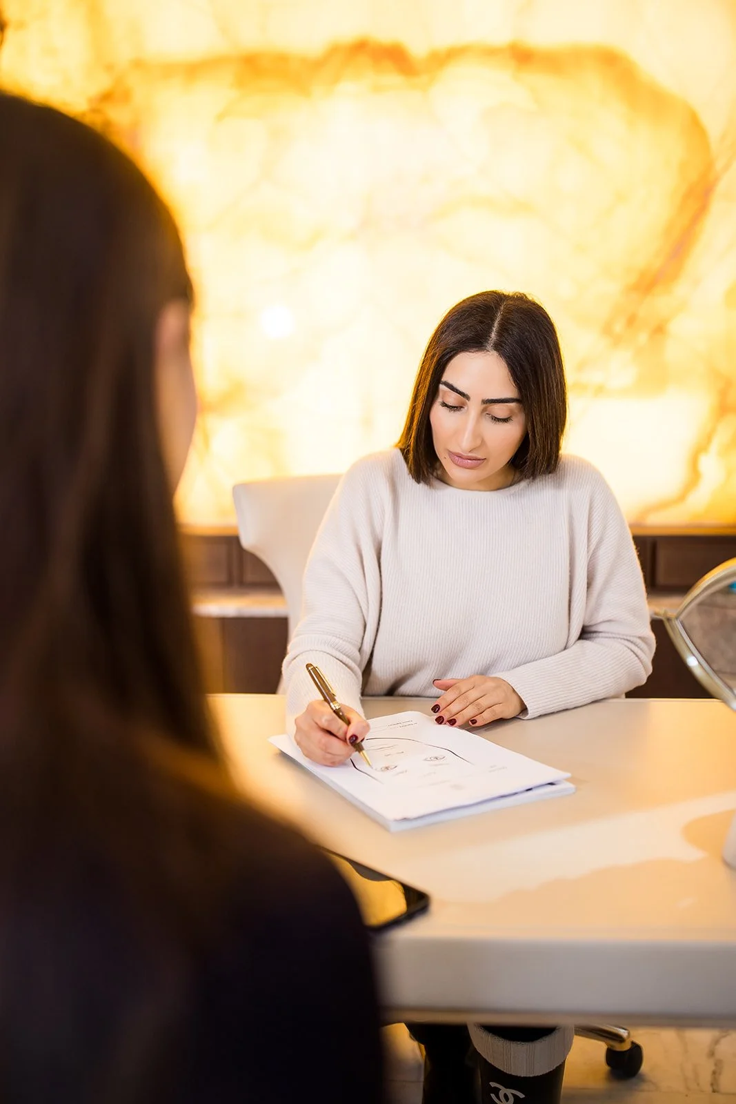 Woman with dark hair in a white sweater sitting at a desk, writing on papers, with a blurred person in black in the foreground, and a warmly lit background with a large illuminated stone wall.