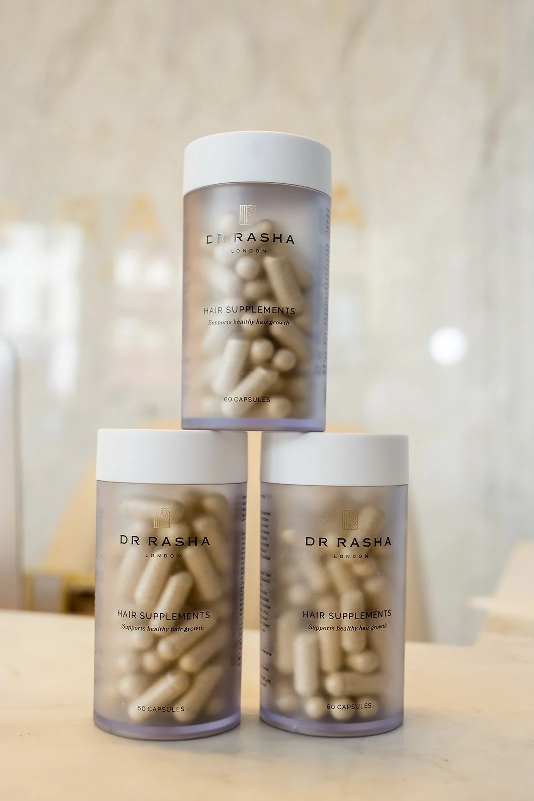 Three containers of Dr Rasha London Hair Supplements with white capsules visible inside, stacked in a pyramid shape on a light-colored surface.